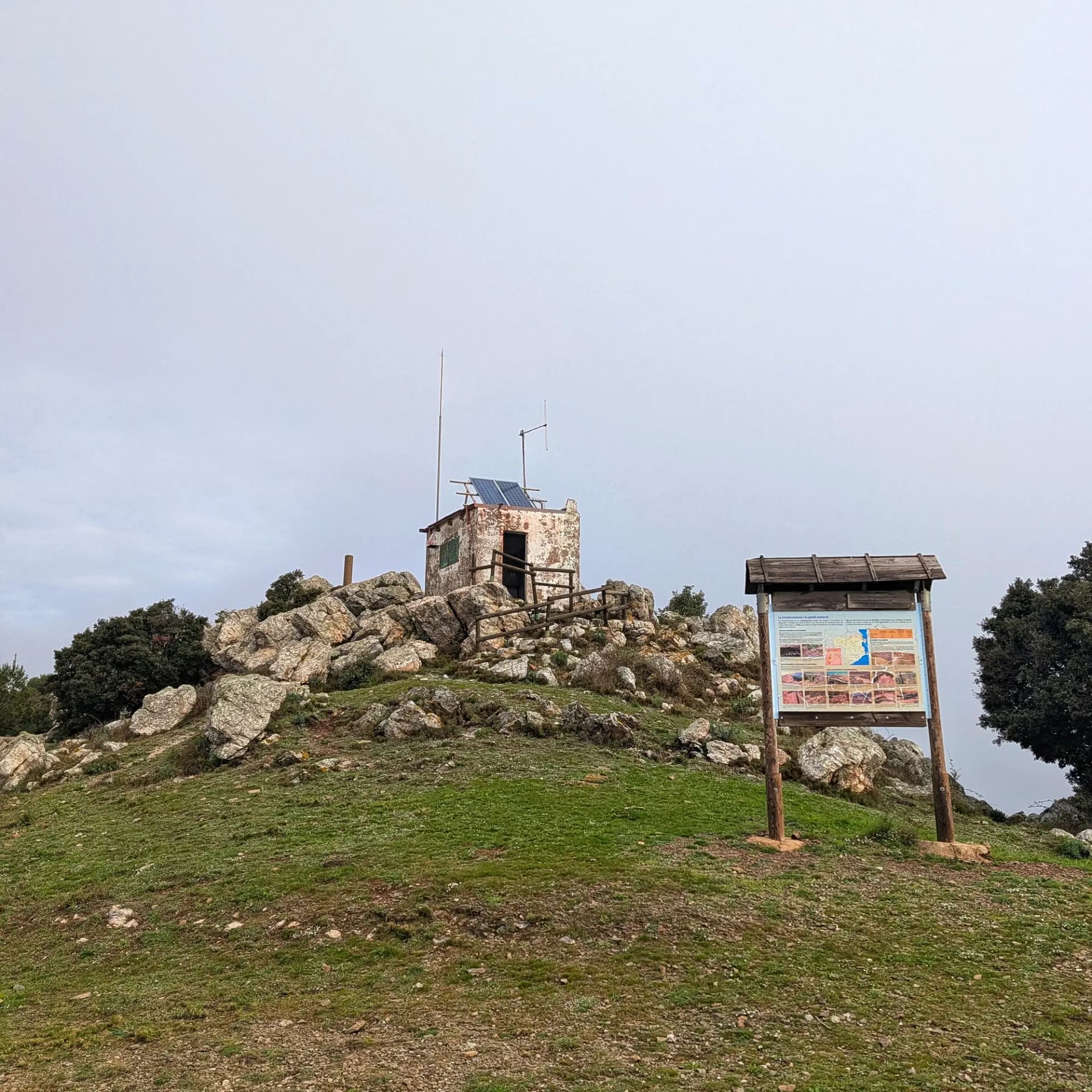 Edifici amb panell solar sobre el cim rocós i herbós del Puig d'Esquers, amb panell informatiu i cel cobert.