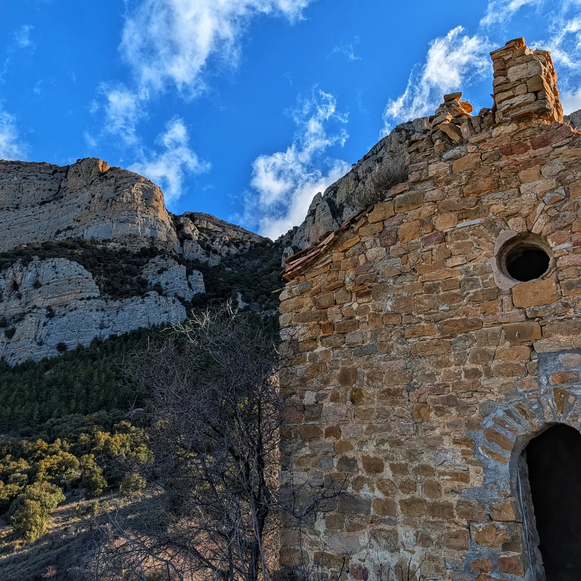 Ruinas de la Ermita de Rúbies, de piedra, con ventana redonda y puerta arqueada, montañas y cielo azul con nubes.
