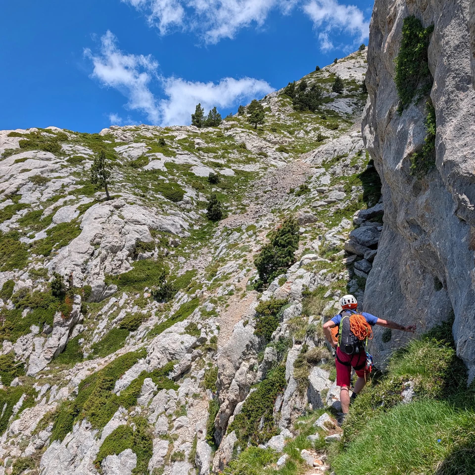Mountaineer with helmet, backpack, and rope on a steep, rocky path with green vegetation on Pedraforca under blue sky.