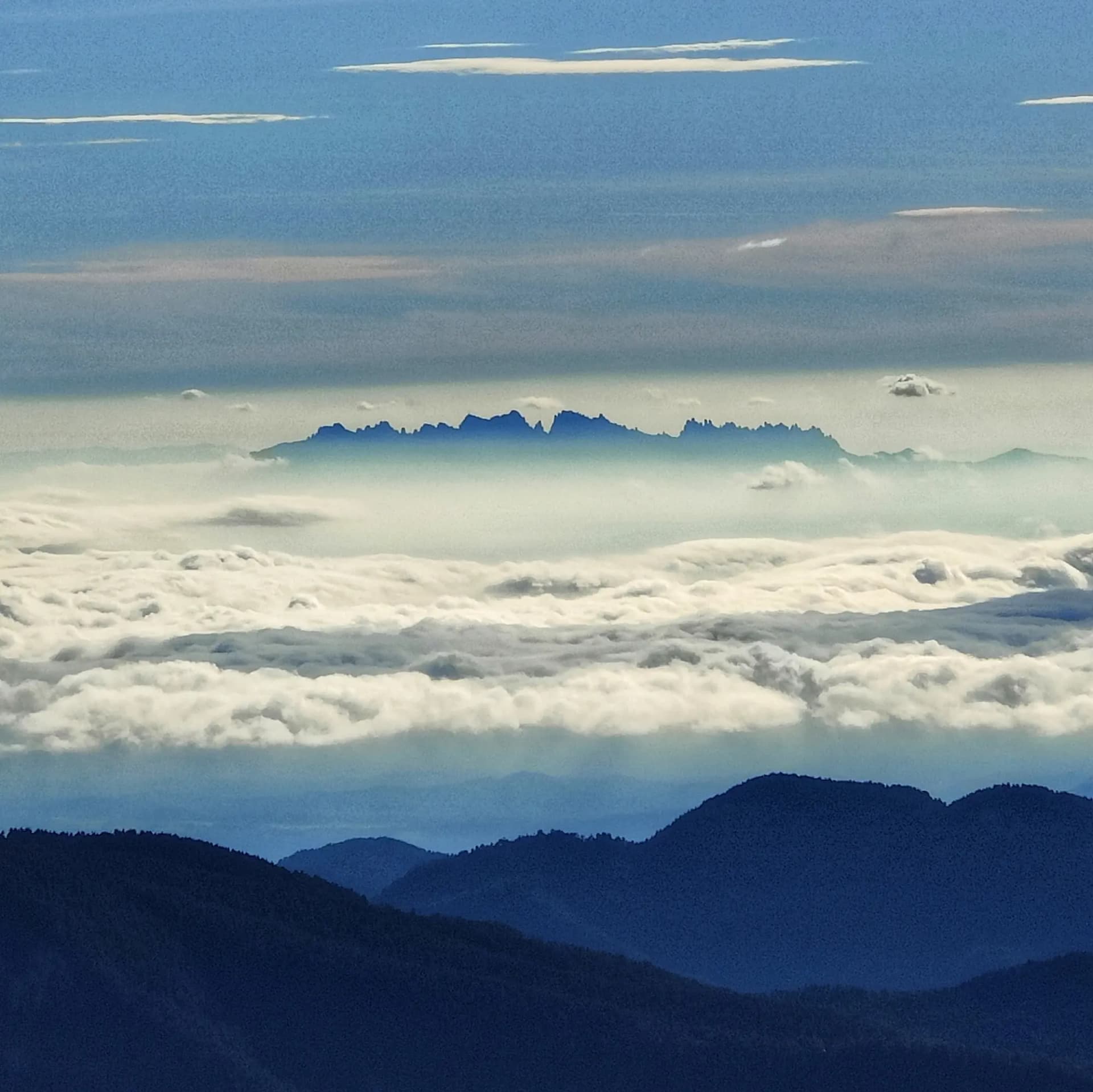 Silueta de Montserrat sobre un mar de nubes, con montañas oscuras en primer plano y cielo claro.