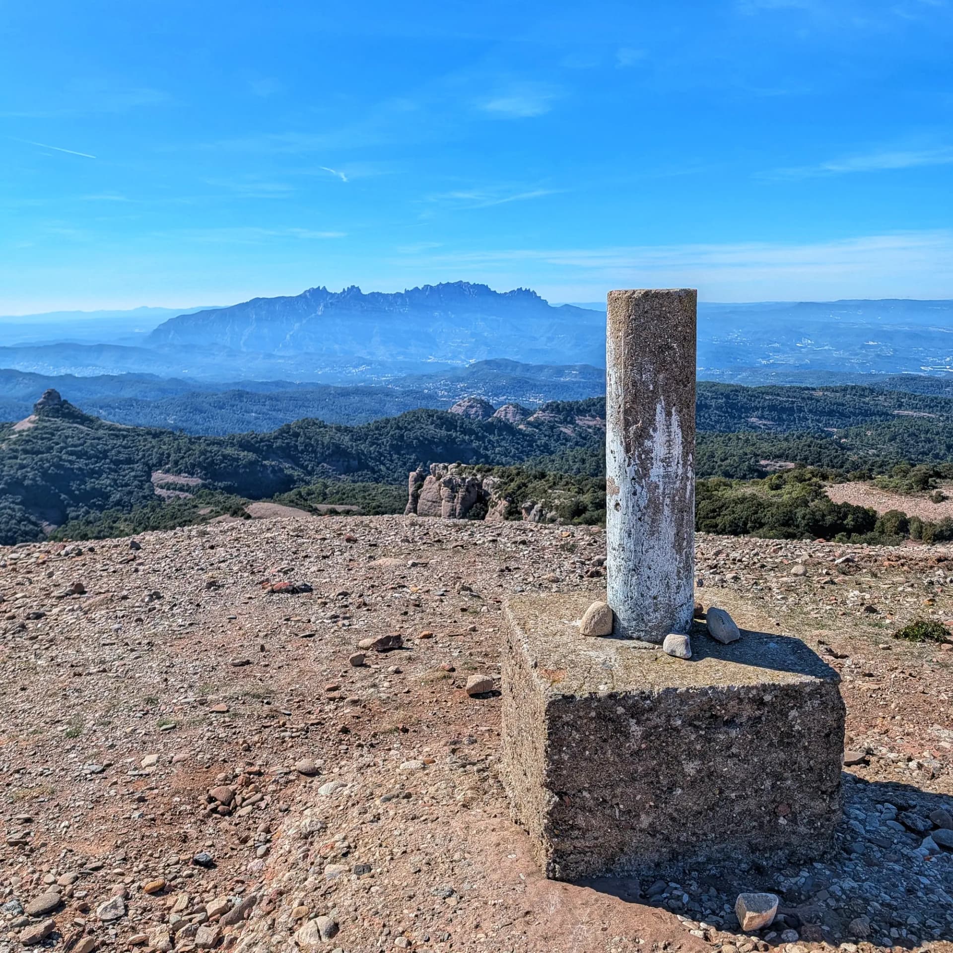 Hito geodésico de hormigón en la cima del Turó de la Pola, con la sierra de Montserrat al fondo bajo cielo azul claro.