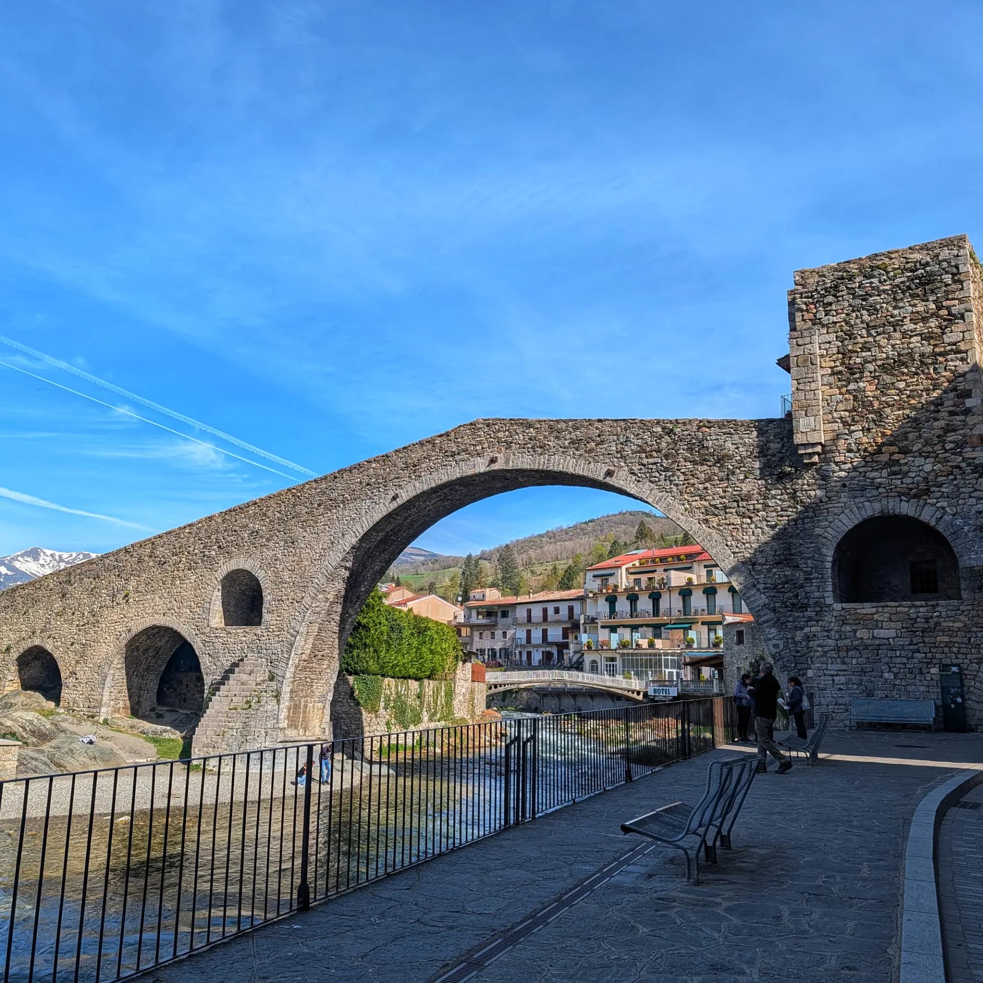 Pont medieval de pedra amb diversos arcs sobre un riu, amb edificis i muntanyes nevades sota un cel blau clar.