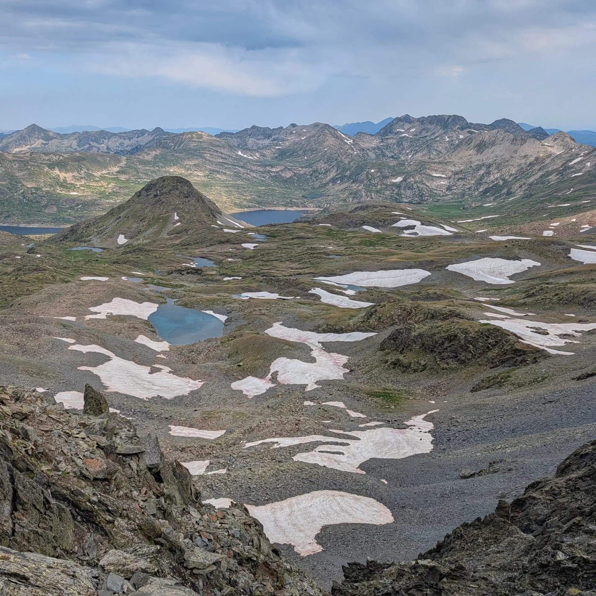 Paisatge alpí amb diversos llacs, clapes de neu blanca sobre terreny rocós i muntanyes distants sota cel ennuvolat.