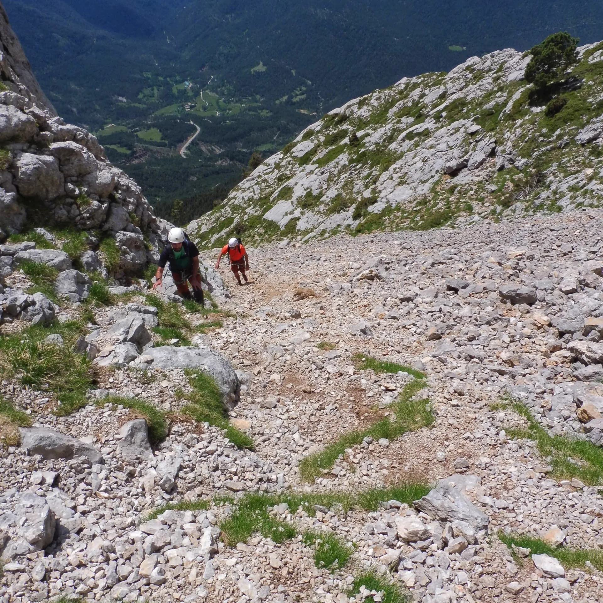 Two climbers ascend a steep rocky scree slope of Pedraforca, with a forested valley and winding road visible below.