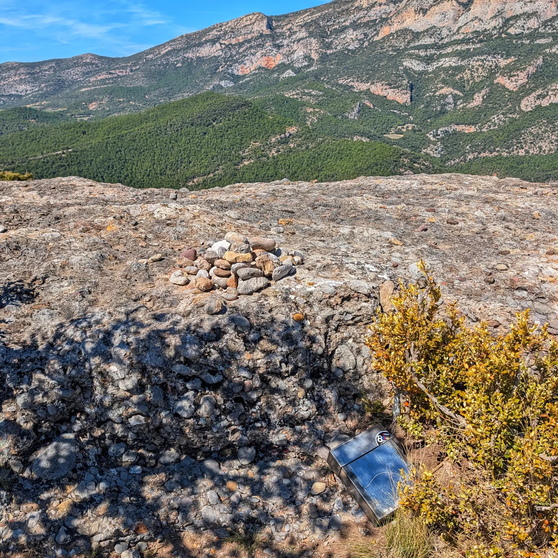Cairn de pedres i bustia metàl·lica a la cimera rocosa de l'Altiplà de Sant Honorat, amb muntanyes boscoses sota cel blau.