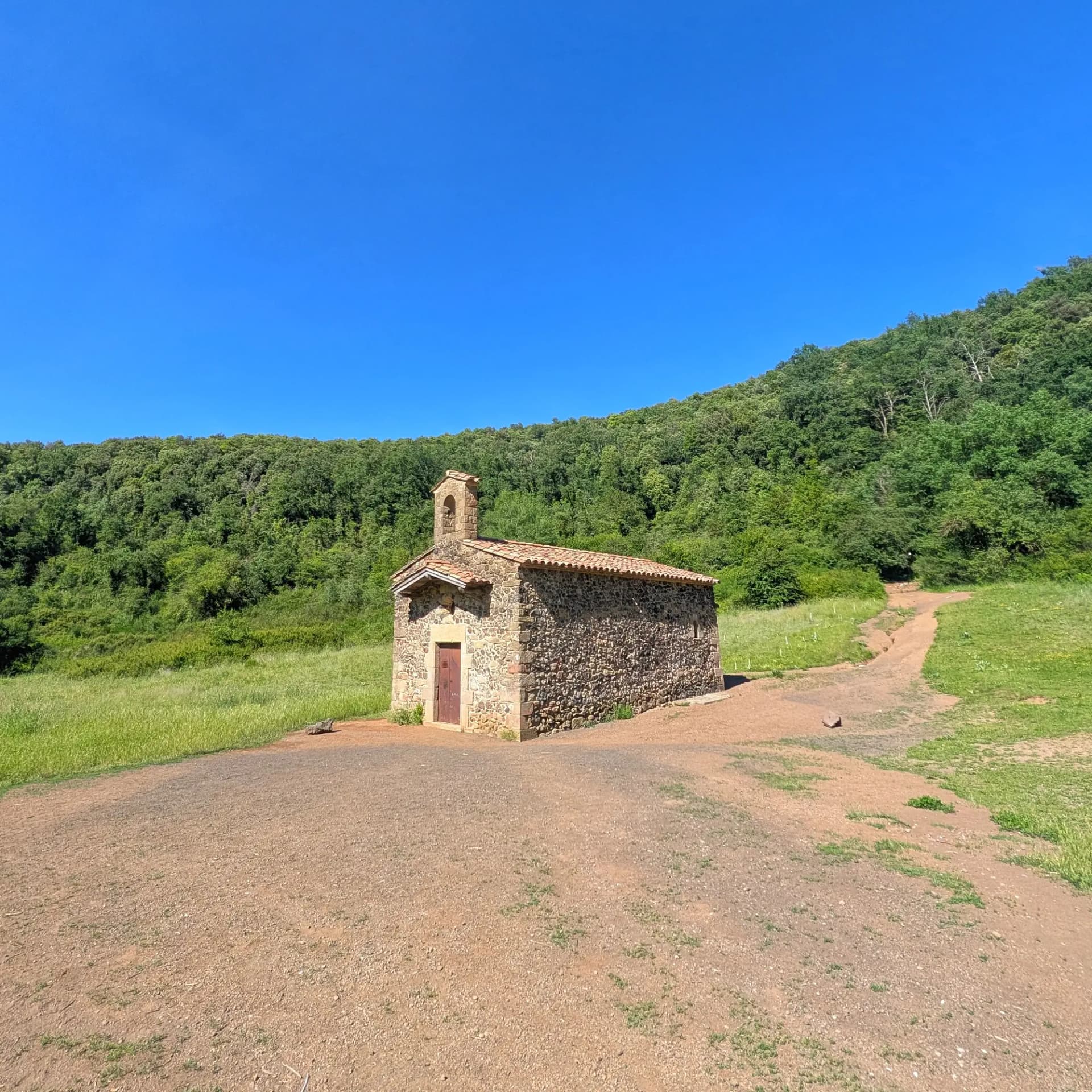 Ermita romànica de pedra amb campana, en una esplanada de terra vermella envoltada de bosc verd sota cel blau.