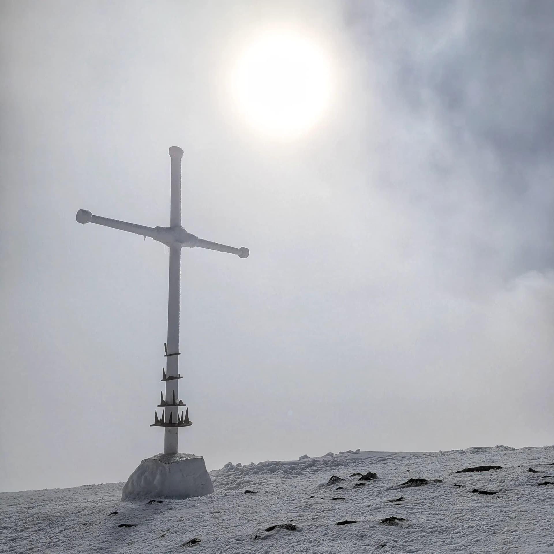 Creu blanca al cim nevat del Taga amb el sol entre la boira.
