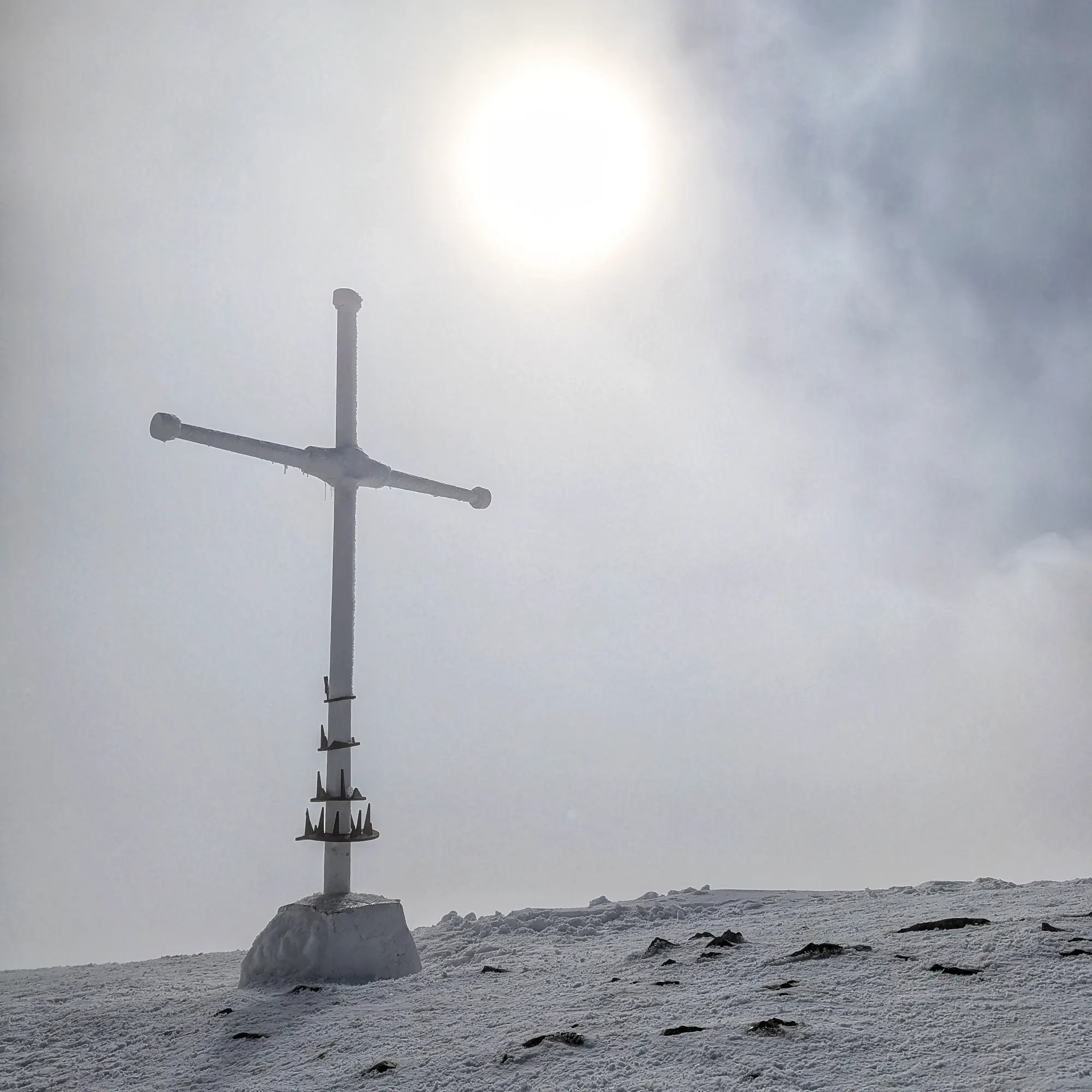 Creu blanca al cim nevat del Taga amb el sol entre la boira.