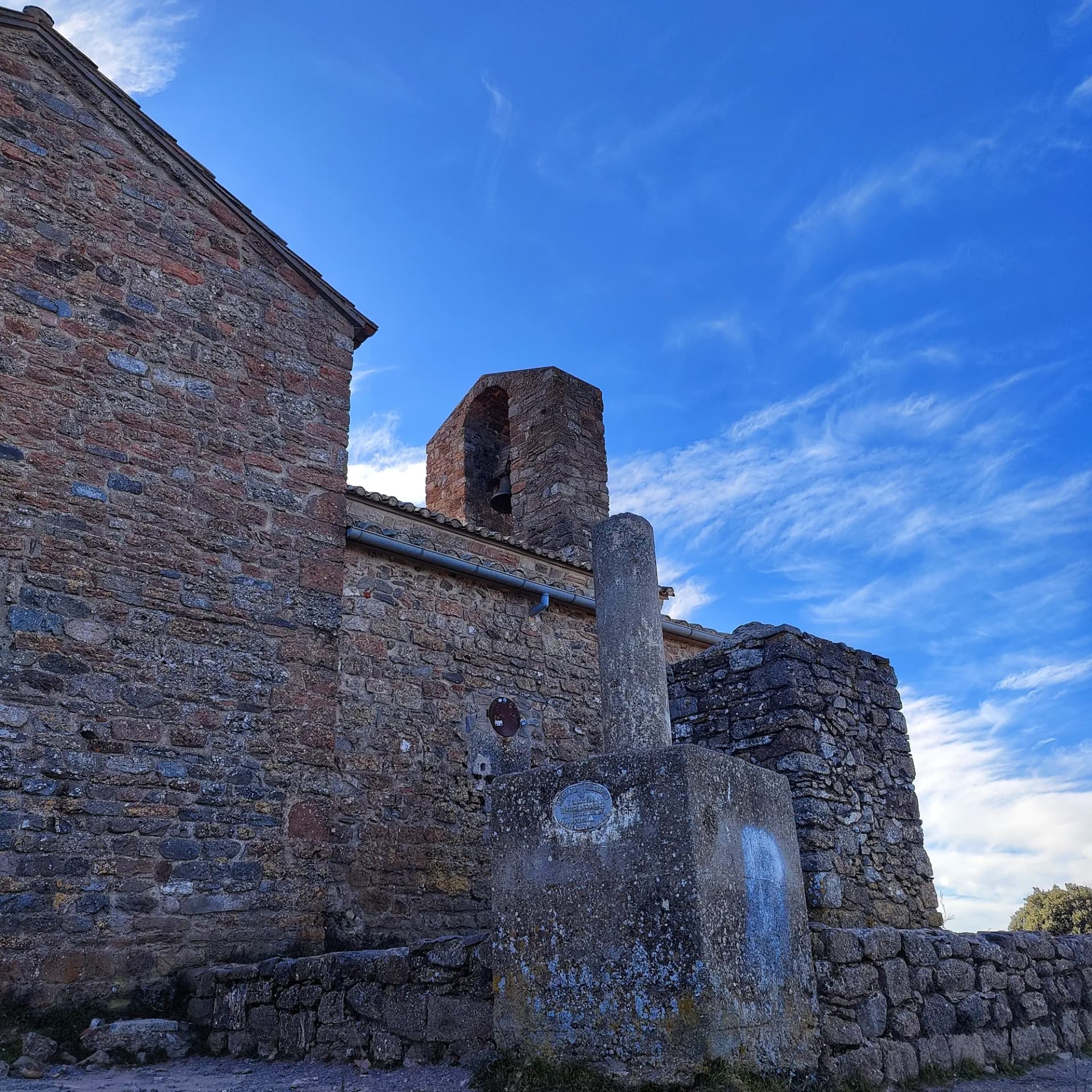 Vista del vèrtex geodèsic de La Mola, emplaçat a Sant Llorenç del Munt, amb l'emblemàtic edifici de pedra del monestir i el seu campanar d'espadanya sota un cel blau amb núvols lleugers. Edifici de pedra amb campanar d'espadanya i vèrtex geodèsic sota cel blau amb núvols lleugers.