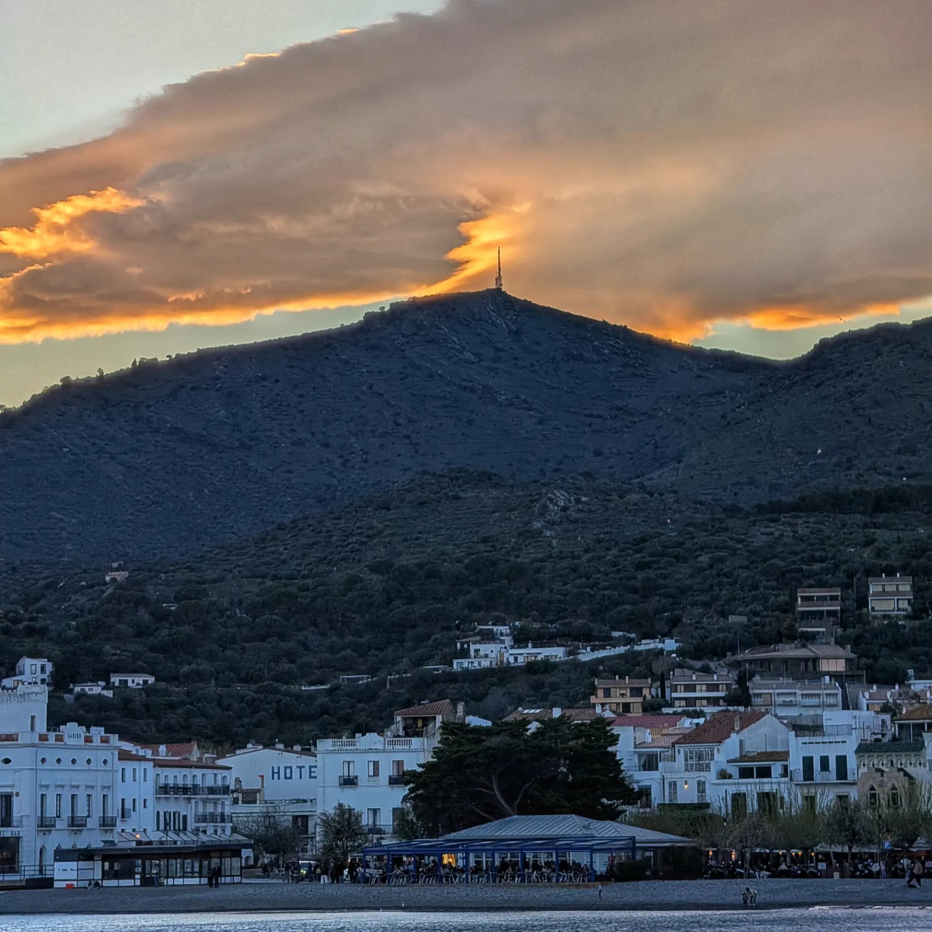 El Puig dels Bufadors amb la seva torre de comunicacions sota un cel de capvespre taronja i gris, vist des de Cadaqués. Al primer pla, edificis blancs del poble i l'activitat a la platja. Puig dels Bufadors amb torre de comunicacions i cel taronja de capvespre sobre Cadaqués, amb edificis i platja.