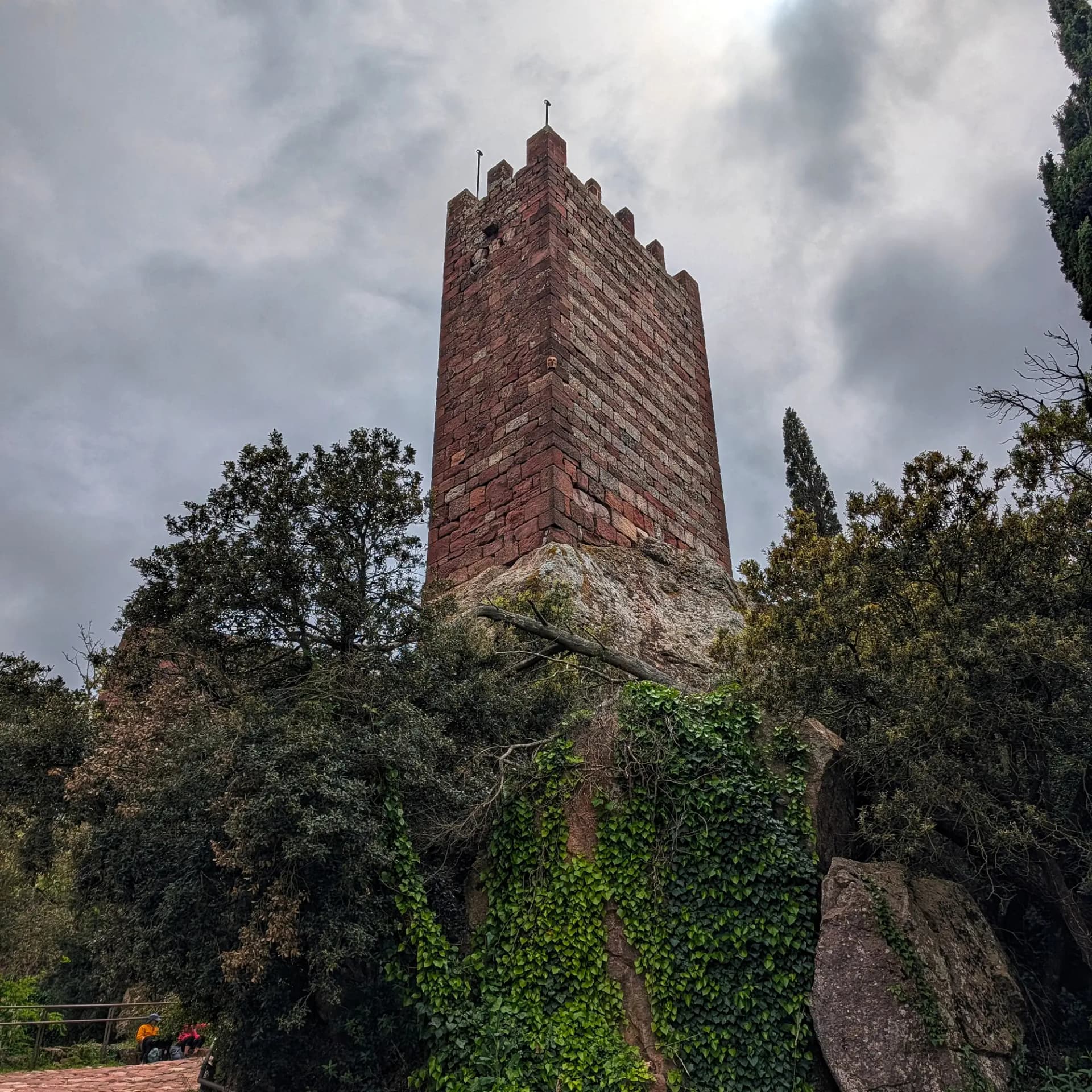 Imatge del Castell d'Escornalbou, mostrant la seva torre principal de tonalitats vermelloses que s'alça sobre una base rocosa i envoltada de vegetació, amb un cel ennuvolat de fons. Torre de pedra vermellosa amb merlets, sobre roques i envoltada de vegetació exuberant sota un cel gris.