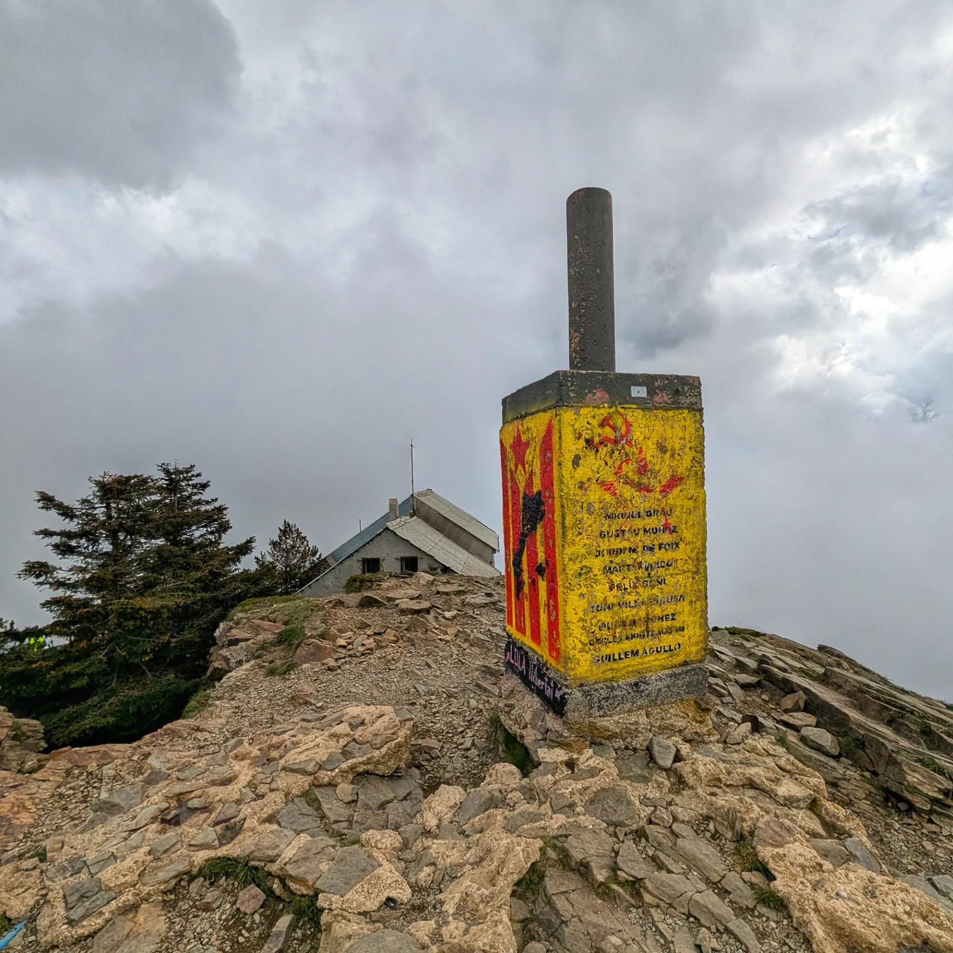 Vista del cim del Turó de l'Home amb la seva fita geodèsica, decorada amb una estelada, i el refugi de muntanya al fons sota un cel cobert. Fita cimera del Turó de l'Home amb estelada esvaïda, refugi de muntanya i cel ennuvolat.