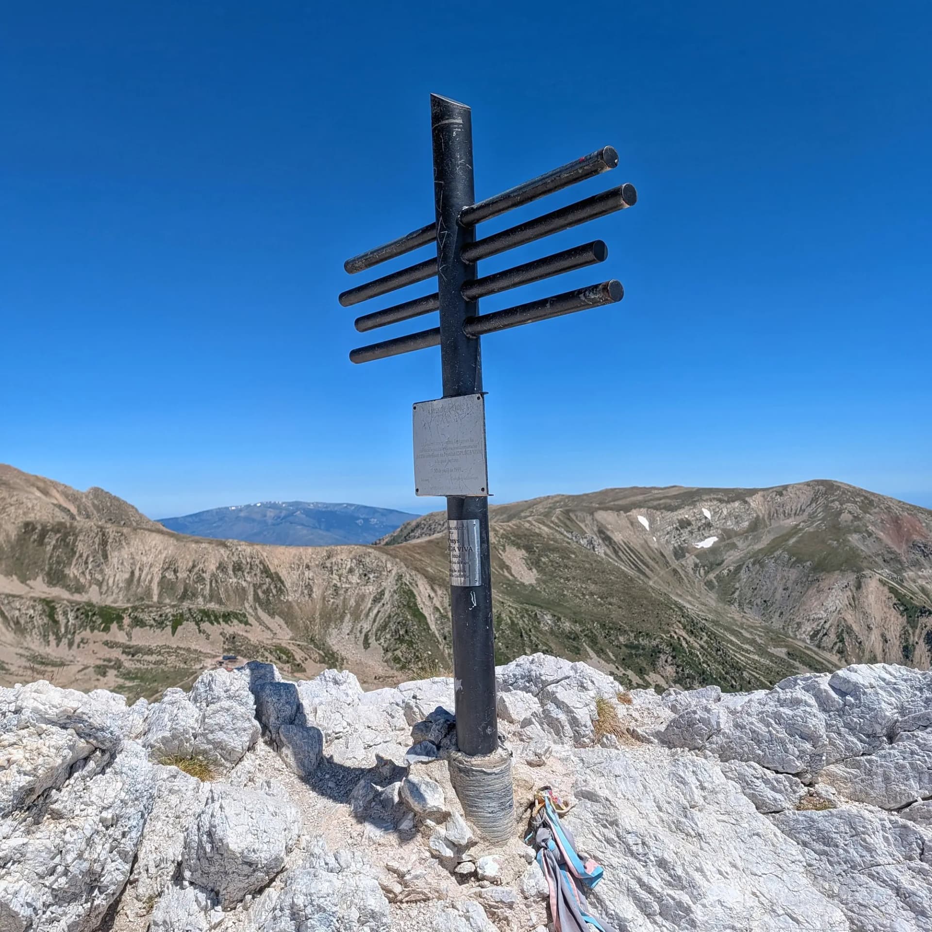Fita de cim al rocós Gra de Fajol, amb vistes panoràmiques a les majestuoses muntanyes dels Pirineus sota un cel blau radiant. Fita metàl·lica fosca sobre un cim rocós sota un cel blau clar, amb pics distants i clapes de neu.
