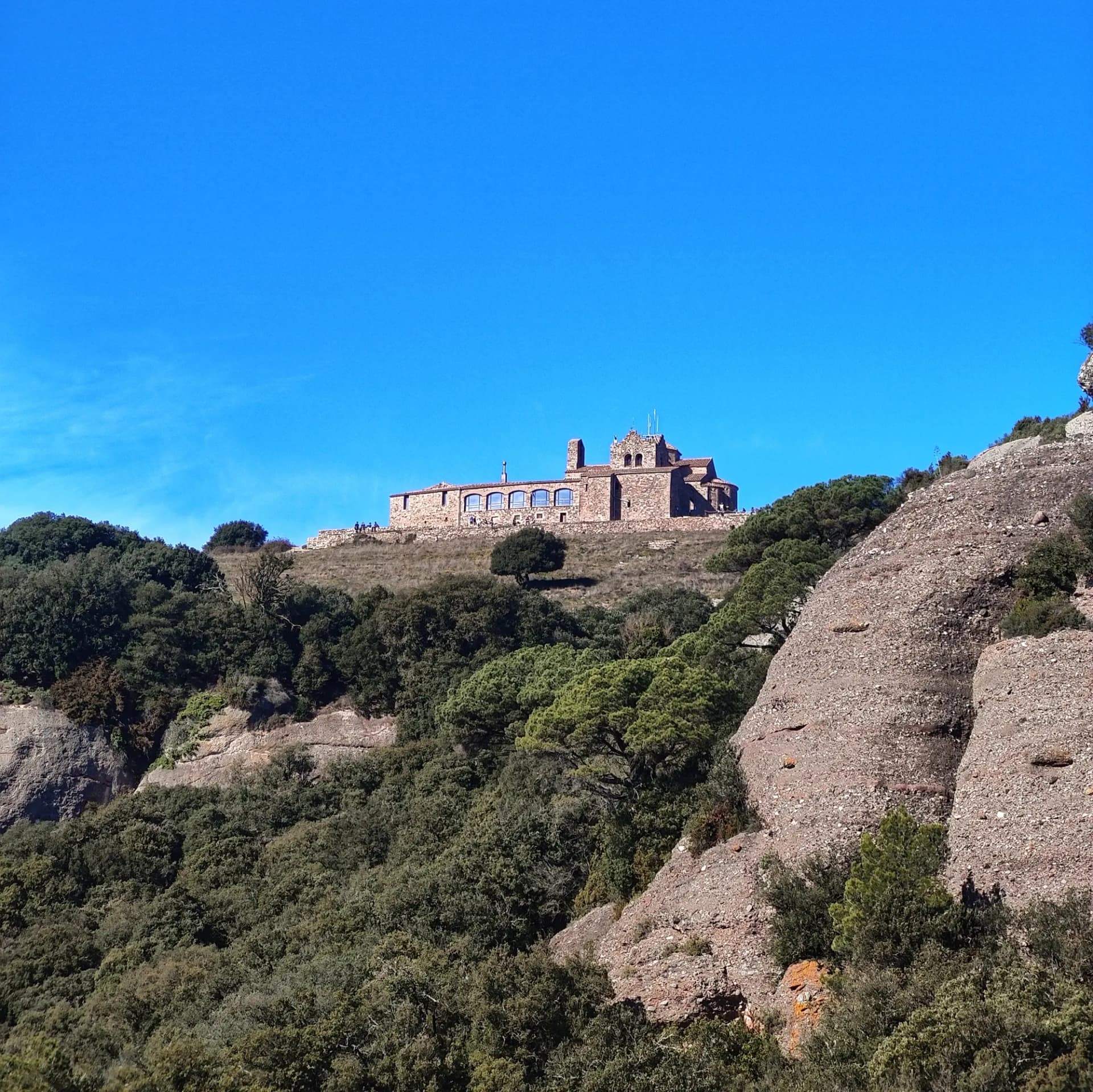 Vista panoràmica del Monestir de Sant Llorenç del Munt, situat estratègicament al cim d'una de les agulles del massís, envoltat per la característica vegetació mediterrània del Parc Natural. Monestir de pedra al cim d'una muntanya, envoltat de bosc i roques, sota un cel blau clar.