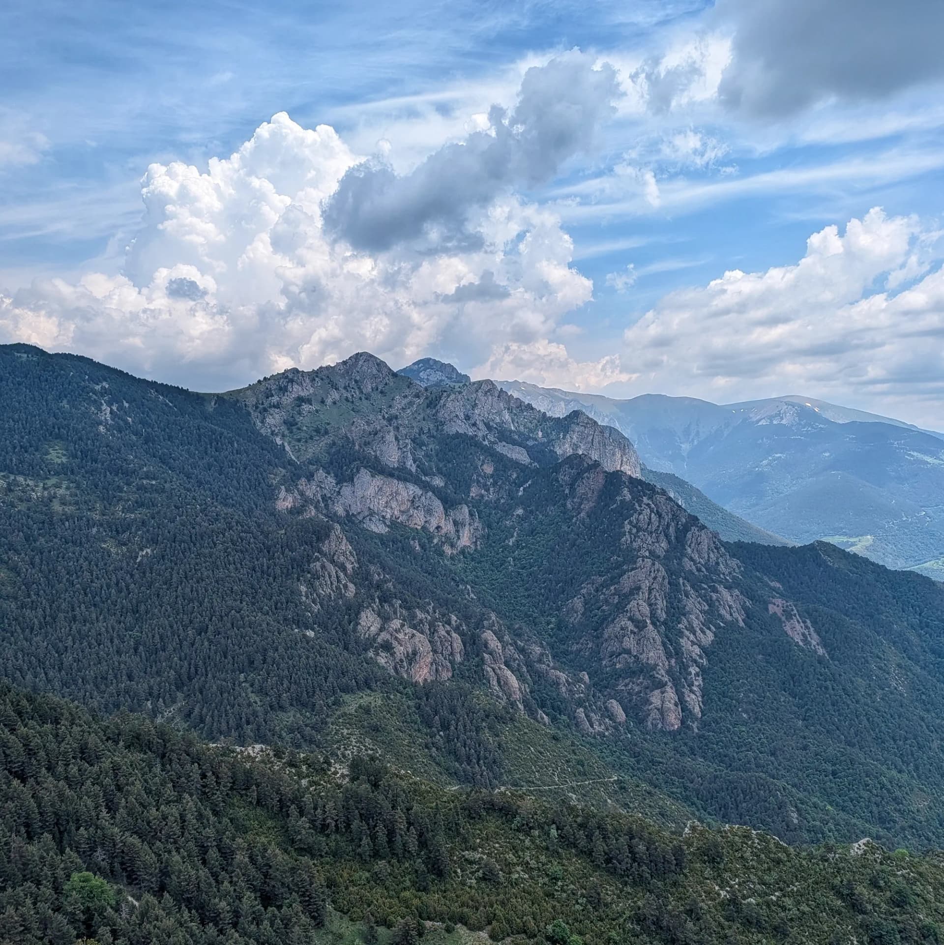 Vista panoràmica cap a les Penyes Altes del Moixeró, mostrant els seus vessants densament boscosos i cims rocosos, observada des del Cap de la Boixassa sota un cel parcialment ennuvolat. Vista panoràmica de muntanyes amb boscos densos, pics rocosos i un cel dinàmic amb núvols blancs.