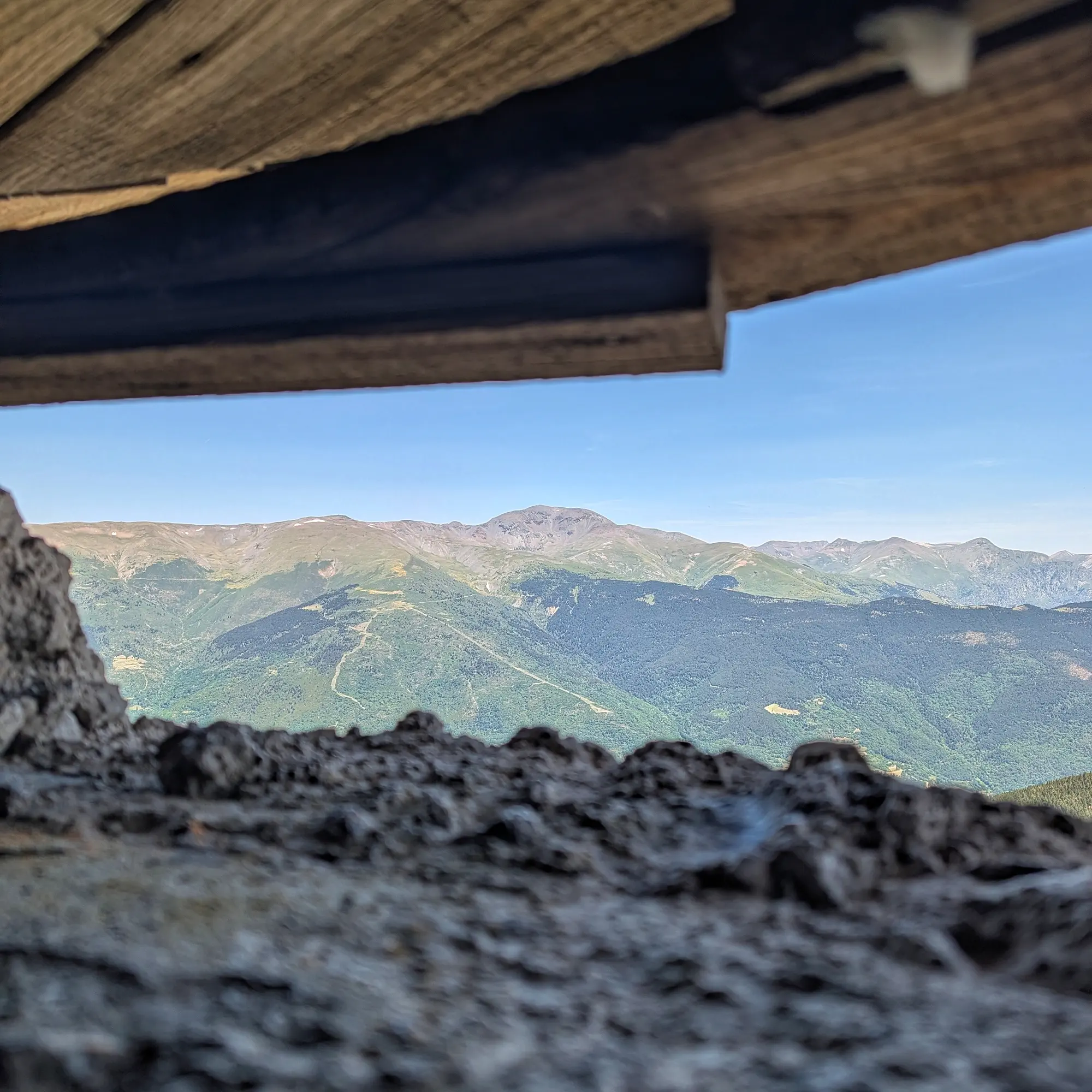 Framed view from the Búnquer del Roc dels Llamps, revealing the majestic layered peaks towards Puigmal under a clear sky. A breathtaking perspective of the Pyrenean landscape. Framed view from a bunker opening: layered green mountains under a clear blue sky, with rough rock and wood.