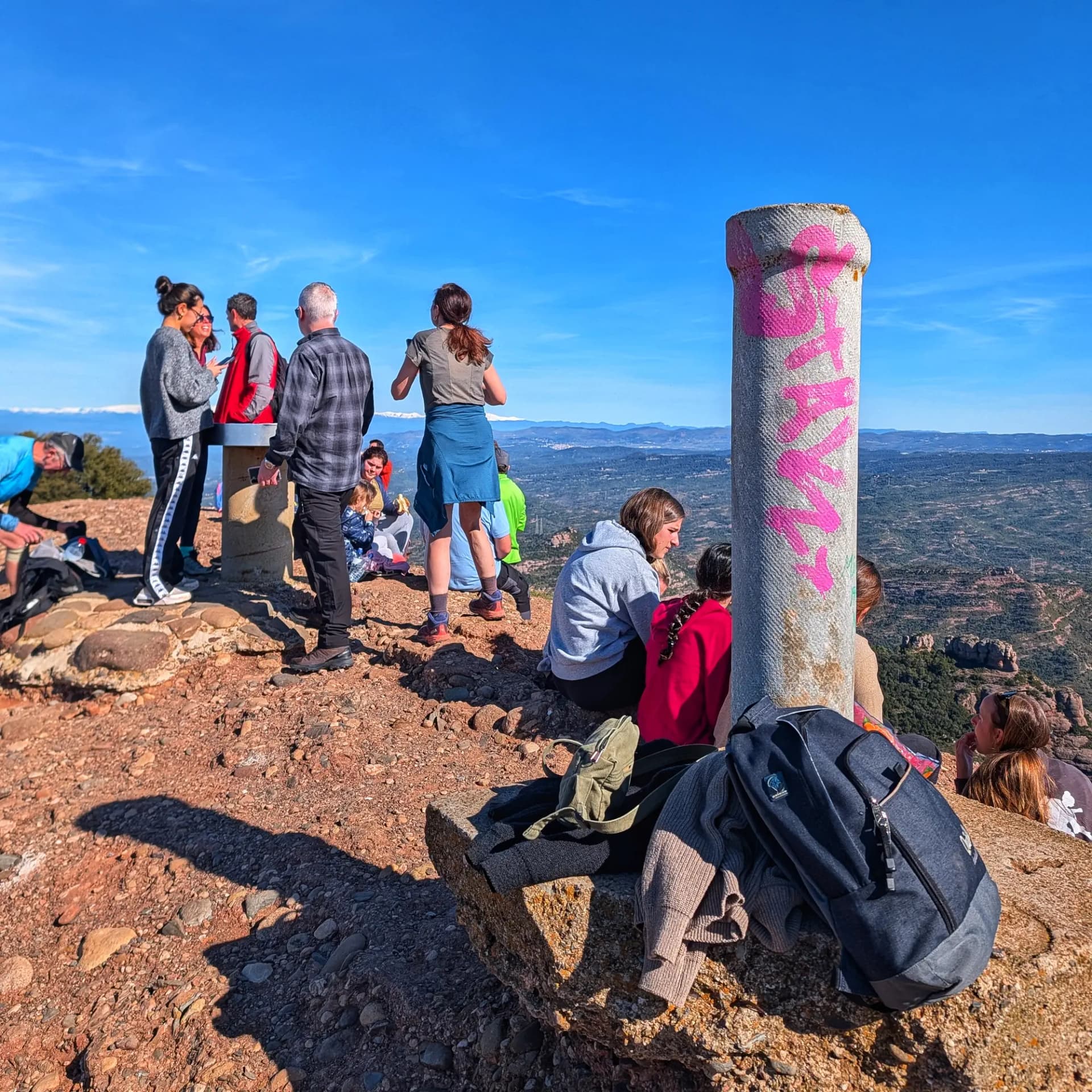 Momento de descanso en la cima del Montcau, durante la ruta por el Parque Natural de Sant Llorenç del Munt i l'Obac que incluye la ascensión a La Mola, Montcau, Turó de la Pola y Castellsapera. Grupo de excursionistas en la cima rocosa del Montcau, con un pilar grafitado y vistas panorámicas bajo cielo azul.