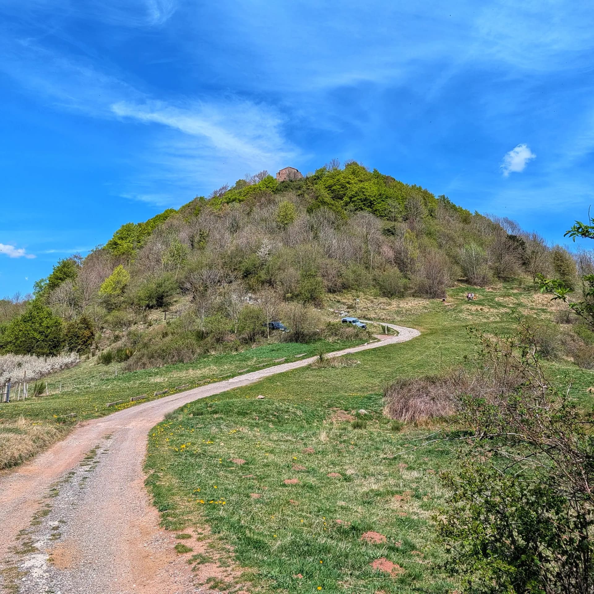 Vista de l'ermita de Sant Antoni a Camprodon, capturada des del Coll de la Brossa, amb un camí de terra que serpenteja turó amunt en un dia assolellat de primavera. Turó verd parcialment boscós amb una ermita de pedra a la cima, un camí de terra sinuós i cel blau amb núvols.