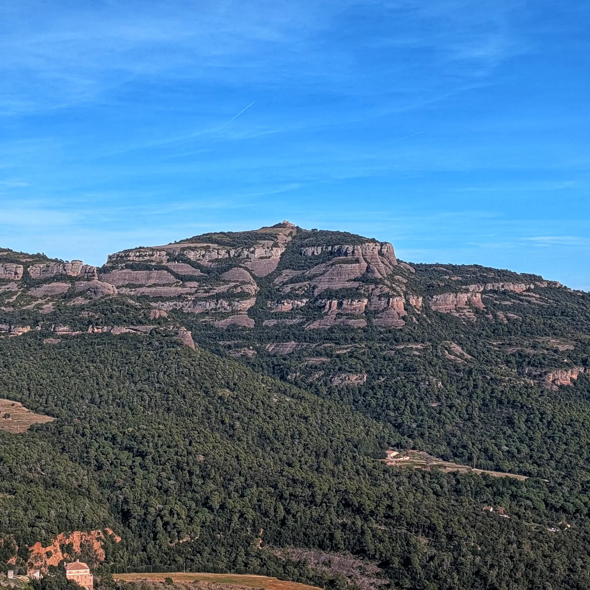 Vista de La Mola, amb el seu monestir al cim, des de la ruta d'ascensió al Parc Natural de Sant Llorenç del Munt i l'Obac. Aquest itinerari inclou l'ascensió a La Mola, Montcau, Turó de la Pola i Castellsapera. Vista panoràmica de La Mola amb el monestir al cim, envoltada de boscos i formacions rocoses sota un cel blau clar.