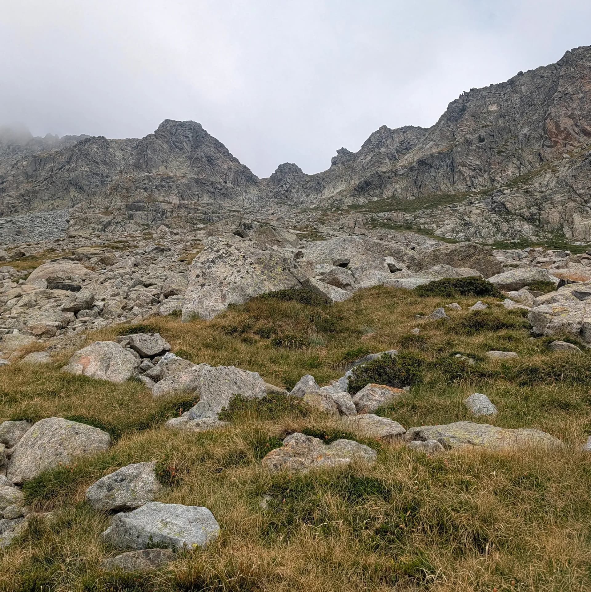 Ascens pel desafiant terreny de granit cap a Comaloforno i Besiberri Sud, des de la Presa de Cavallers. La ruta presenta tarteres extenses i flora de muntanya resistent. Paisatge muntanyós amb grans blocs de granit, herba seca i formacions rocoses escarpades sota cel ennuvolat.