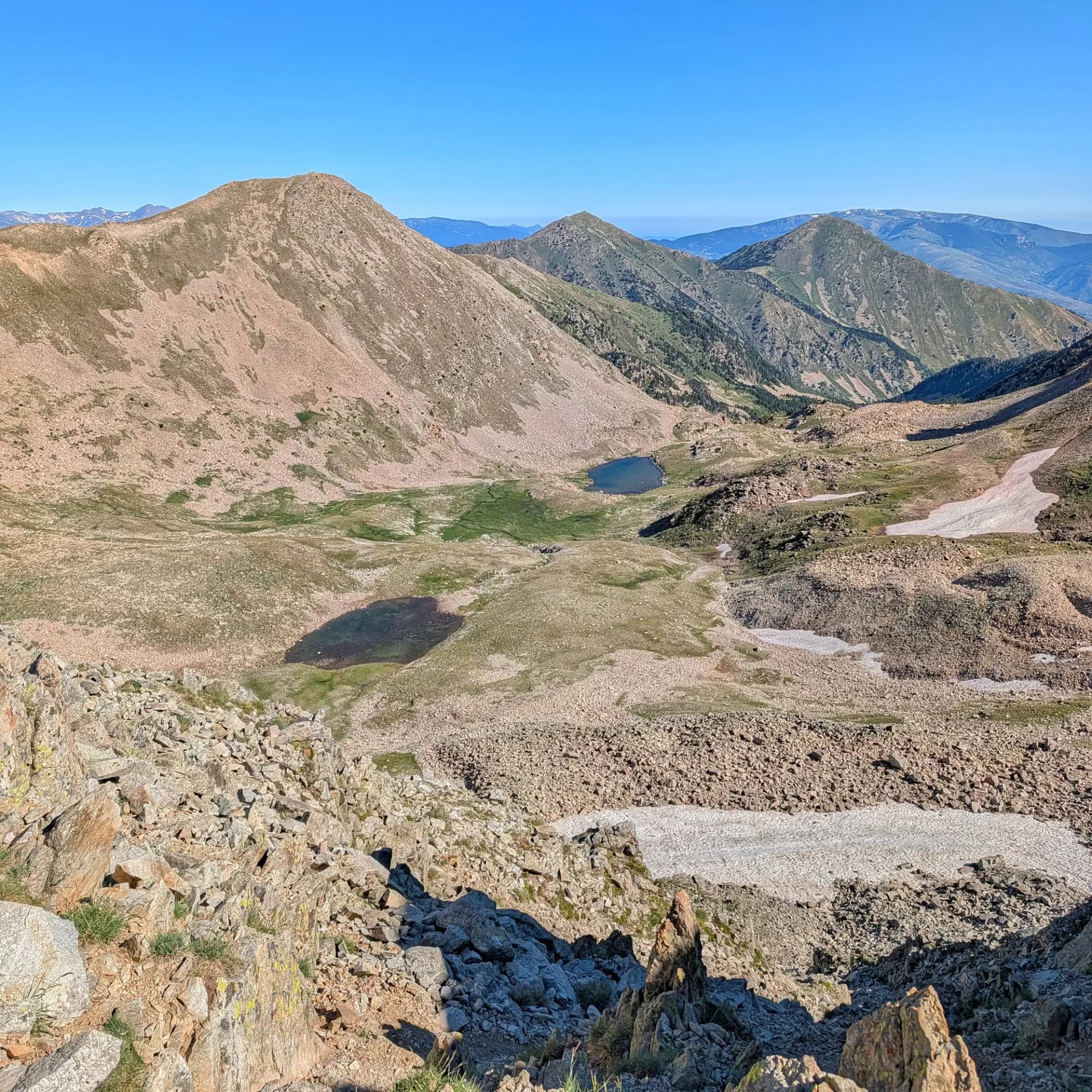 Vista hacia la majestuosa cima de Bastiments (2881m) desde cerca del Coll de Freser, mostrando el paisaje pirenaico con lagos alpinos y parches de nieve. Extenso valle montañoso rocoso con dos pequeños lagos azules, vegetación verde dispersa y parches de nieve bajo un cielo claro.