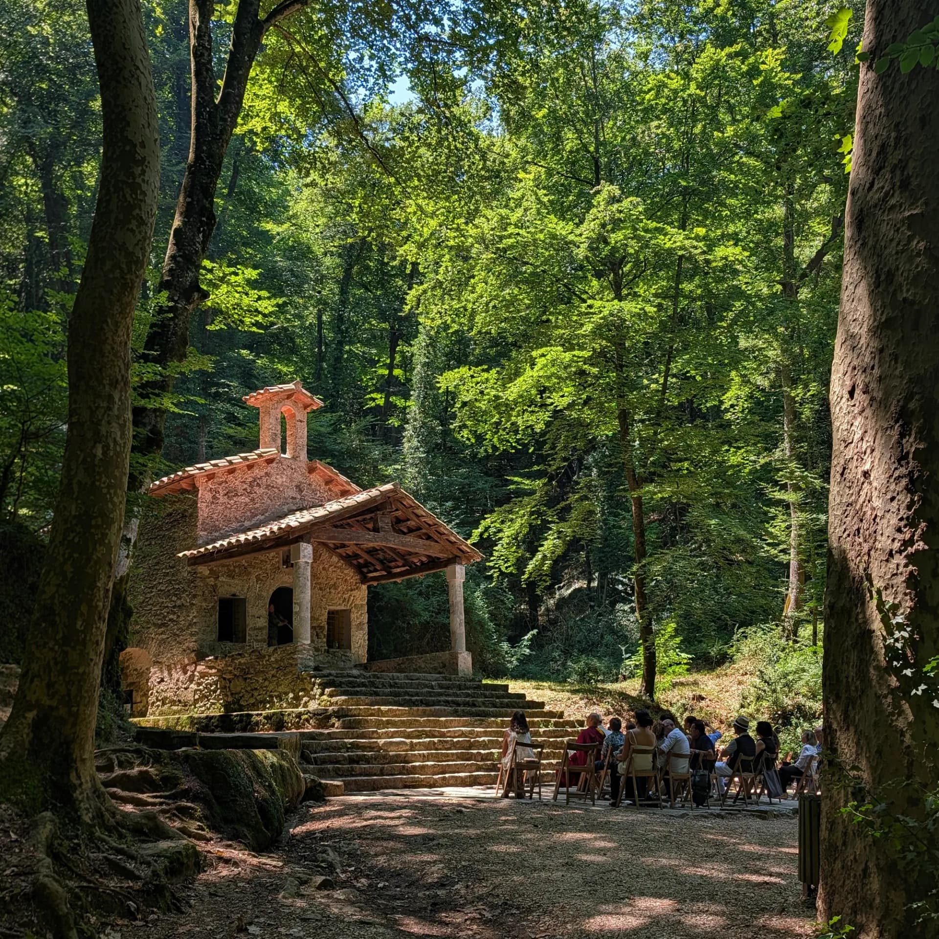 La Ermita de Sant Martí del Corb, una hermosa ermita de piedra, enclavada en un frondoso bosque, acoge un encuentro al aire libre. Ermita de piedra con espadaña en bosque denso y verde, iluminada por el sol. Gente sentada frente a las escaleras.