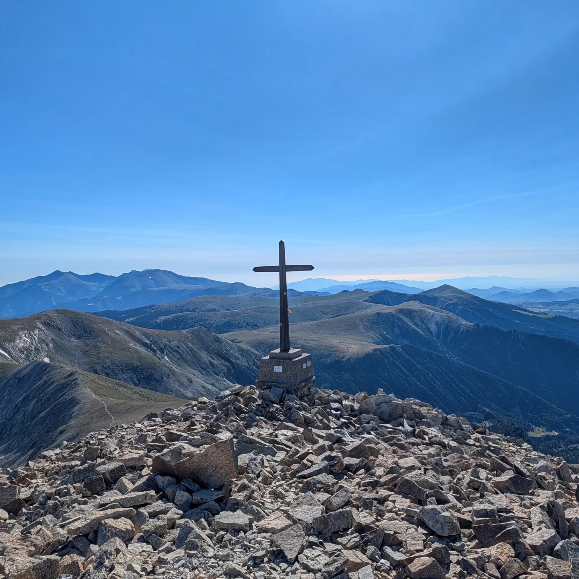 Coronando el Bastiments (2881m), un pico popular que ofrece impresionantes vistas panorámicas de los Pirineos y un atisbo lejano del mar bajo un cielo azul claro. Excursionistas en una cima rocosa con una gran cruz oscura. Amplio panorama de montañas azules lejanas bajo un cielo claro.
