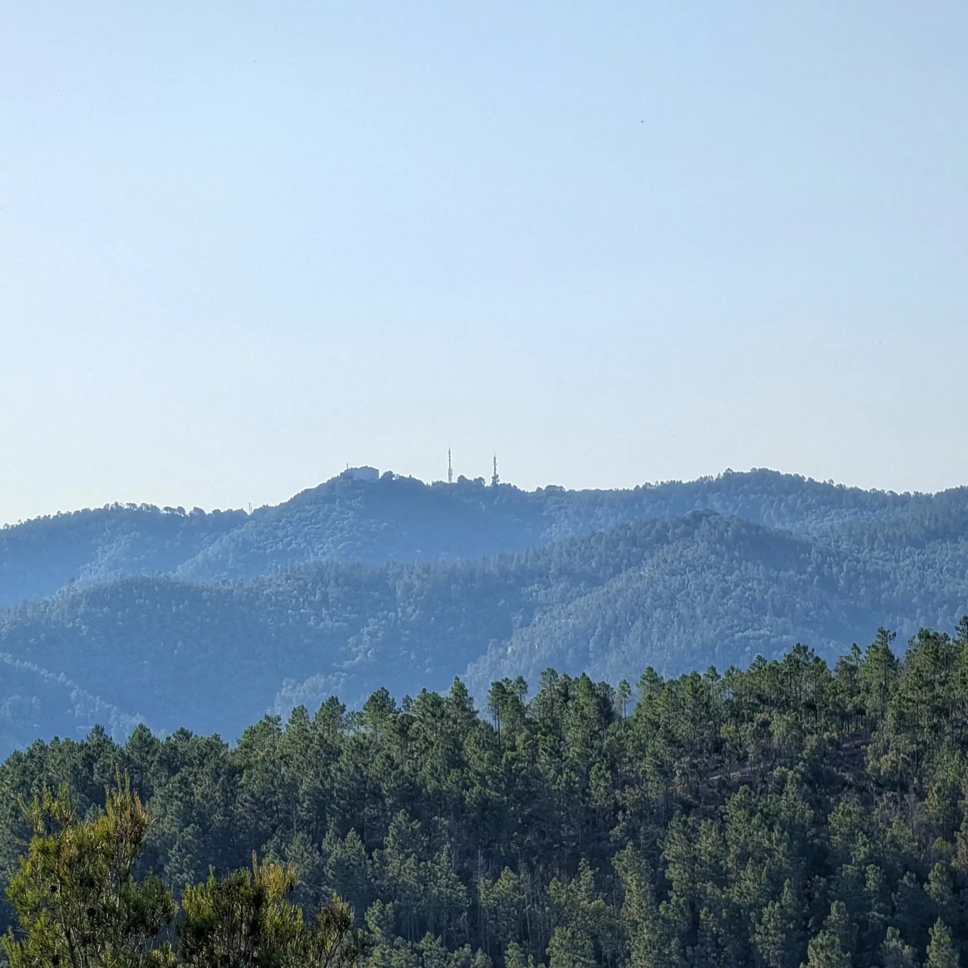 Una vista en capas del paisaje boscoso, presentando la cima de la Muntanya dels Àngels (483m) con su edificio y torres icónicas. Capturado desde el Castillo de Sant Miquel. Paisaje de montañas boscosas en capas bajo cielo azul claro, con edificio y torres de comunicación en la cima lejana.