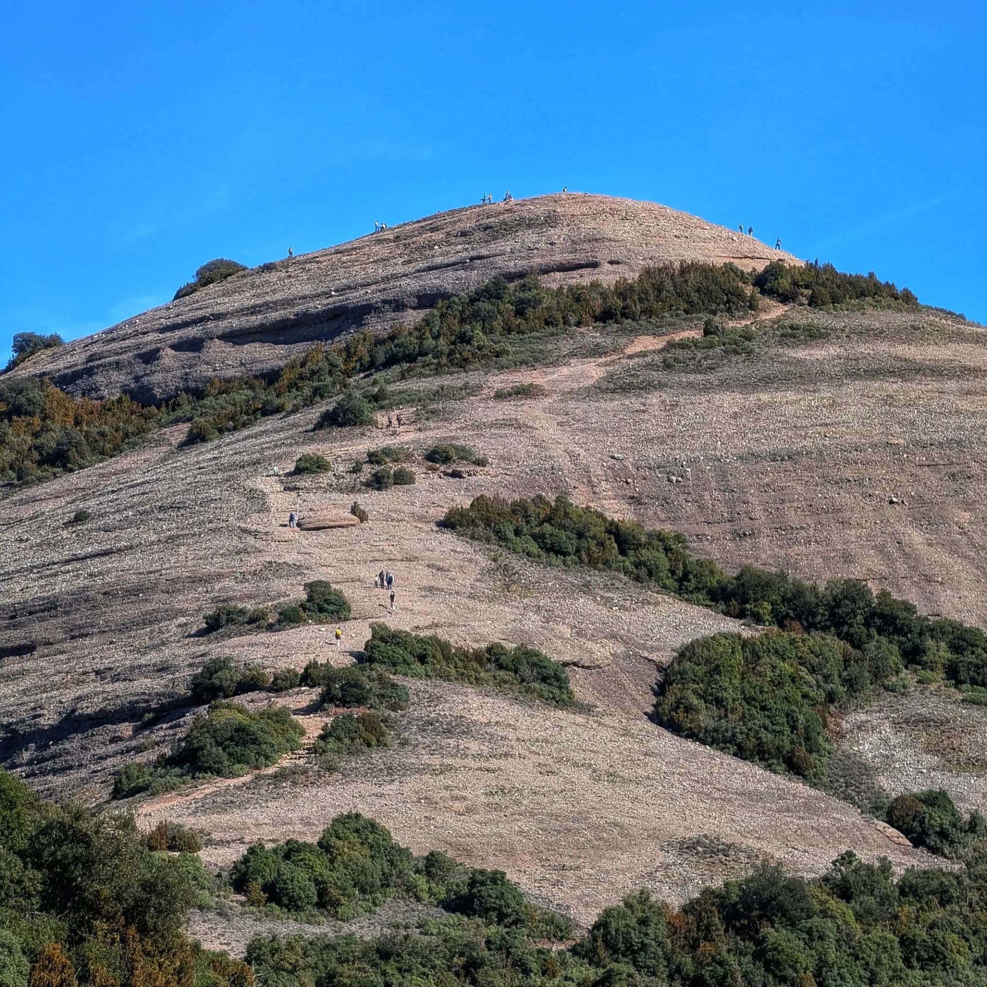 Vistes del Montcau amb els seus característics senders, durant una ruta pel Parc Natural de Sant Llorenç del Munt i l'Obac. Es poden observar diversos grups de senderistes ascendint la muntanya sota un cel clar. Muntanya rocosa Montcau amb senders, petits grups de senderistes i vegetació sota cel blau clar.