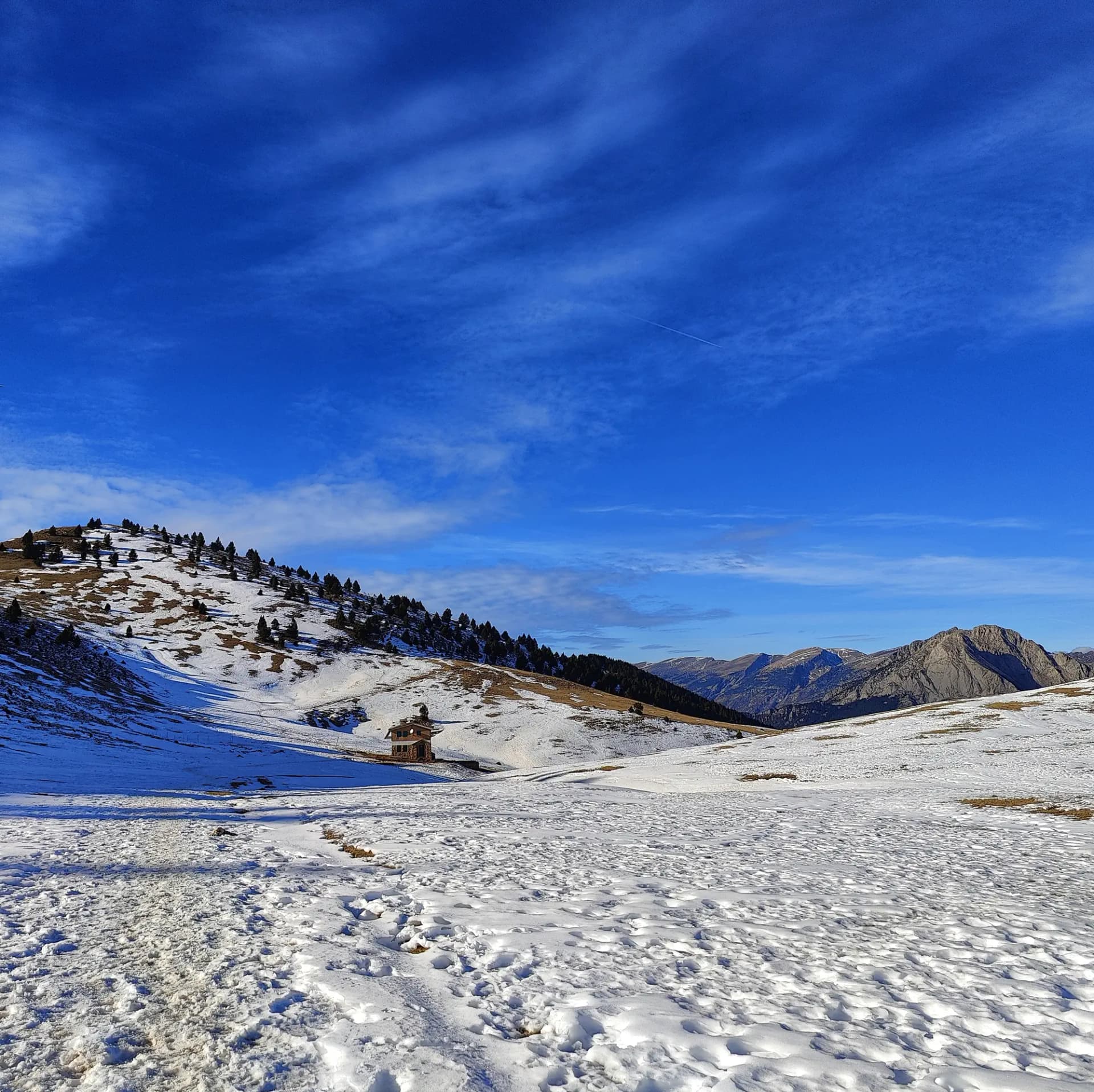 Vista desde la Serra d'Ensija ascendiendo al Cap de la Gallina Pelada, con el pico a la izquierda, el refugio en el centro y el macizo del Pedraforca a la derecha, bajo un cielo azul intenso y con alguna nube dispersa. Paisaje invernal en Serra d'Ensija: refugio en valle nevado, Cap de la Gallina Pelada izquierda, Pedraforca derecha, cielo azul claro.