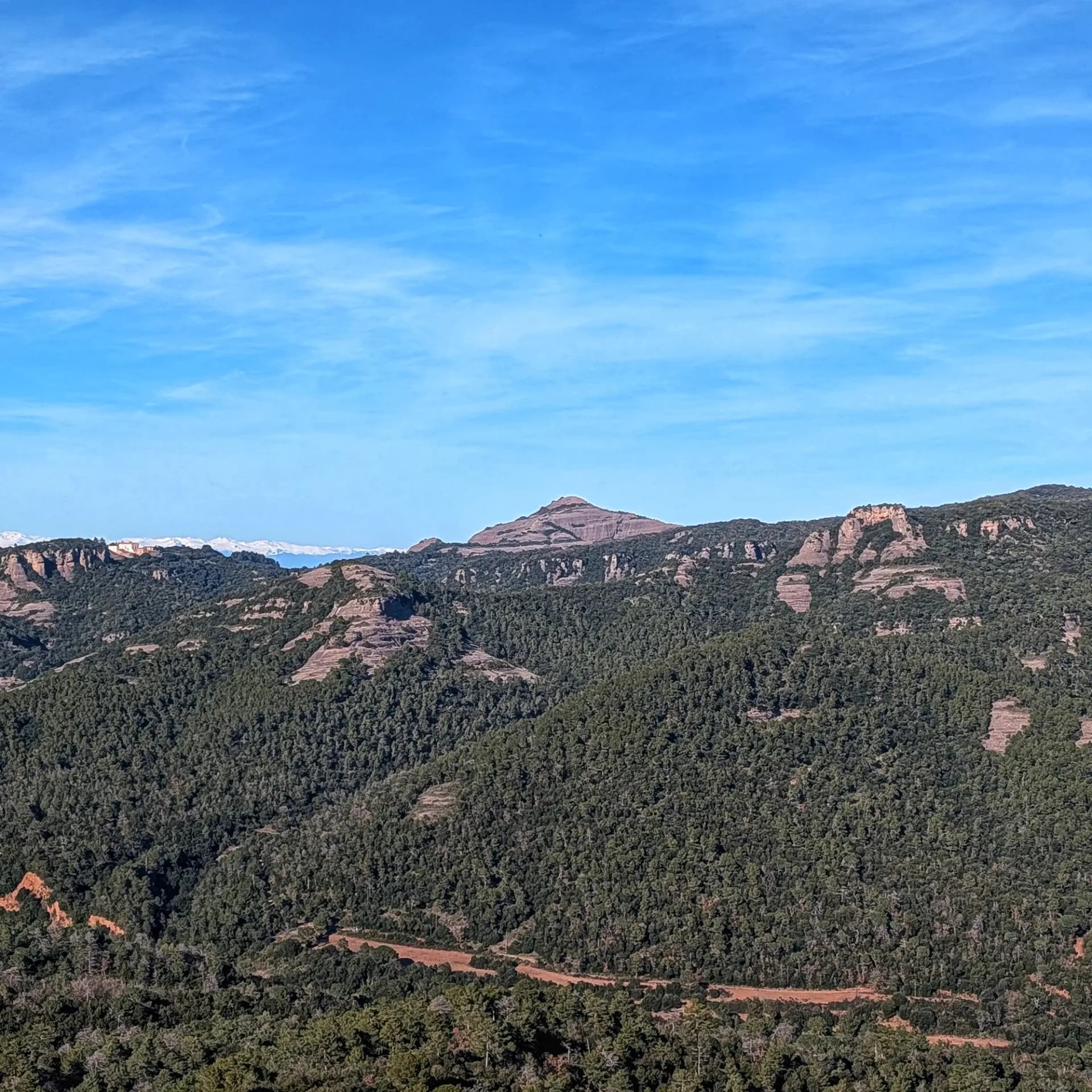 Vistas del Montcau desde la ruta de ascenso a La Mola, en el Parque Natural de Sant Llorenç del Munt i l'Obac. La imagen captura el icónico Montcau, las sierras boscosas, formaciones rocosas y un sendero que serpentea por el paisaje, con cumbres nevadas lejanas bajo un cielo claro. Vistas del Montcau y montañas boscosas bajo un cielo azul en el Parque Natural de Sant Llorenç del Munt i l'Obac.