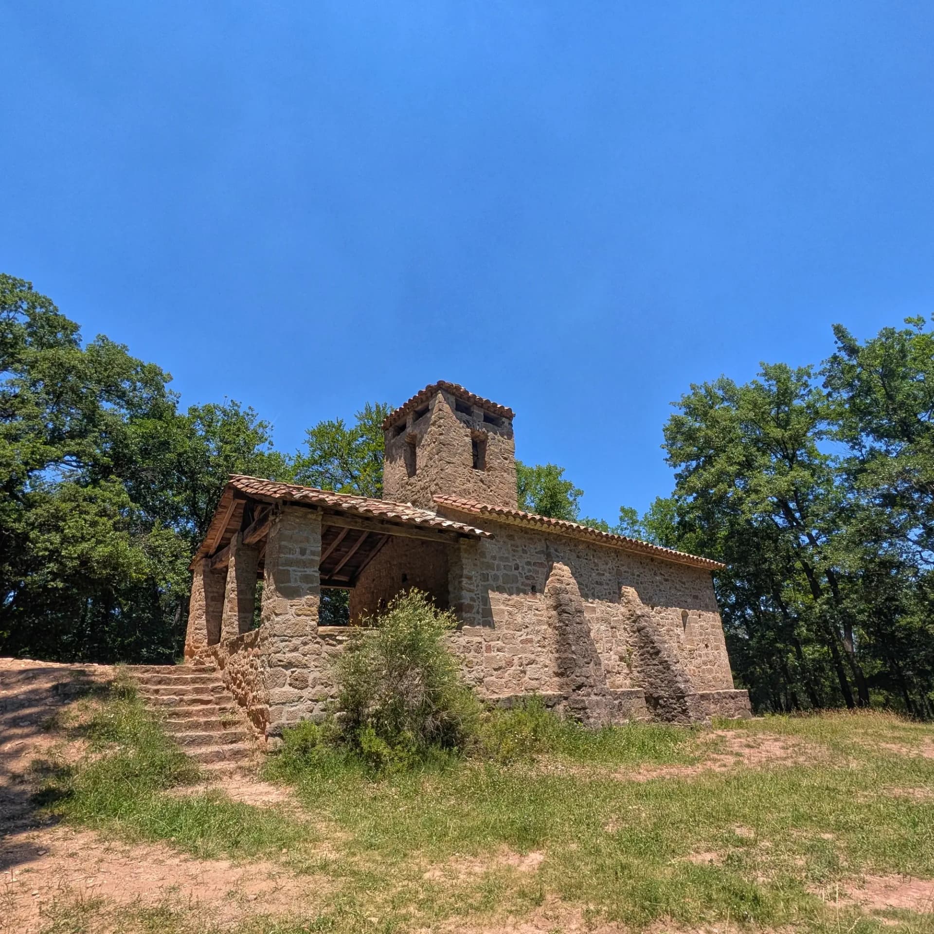 La histórica Ermita de Sant Miquel del Corb, una hermosa ermita de piedra enclavada entre la vegetación bajo un cielo azul radiante. Un rincón de paz. Ermita de piedra con tejado de tejas y torre, rodeada de árboles verdes y hierba bajo un cielo azul claro.