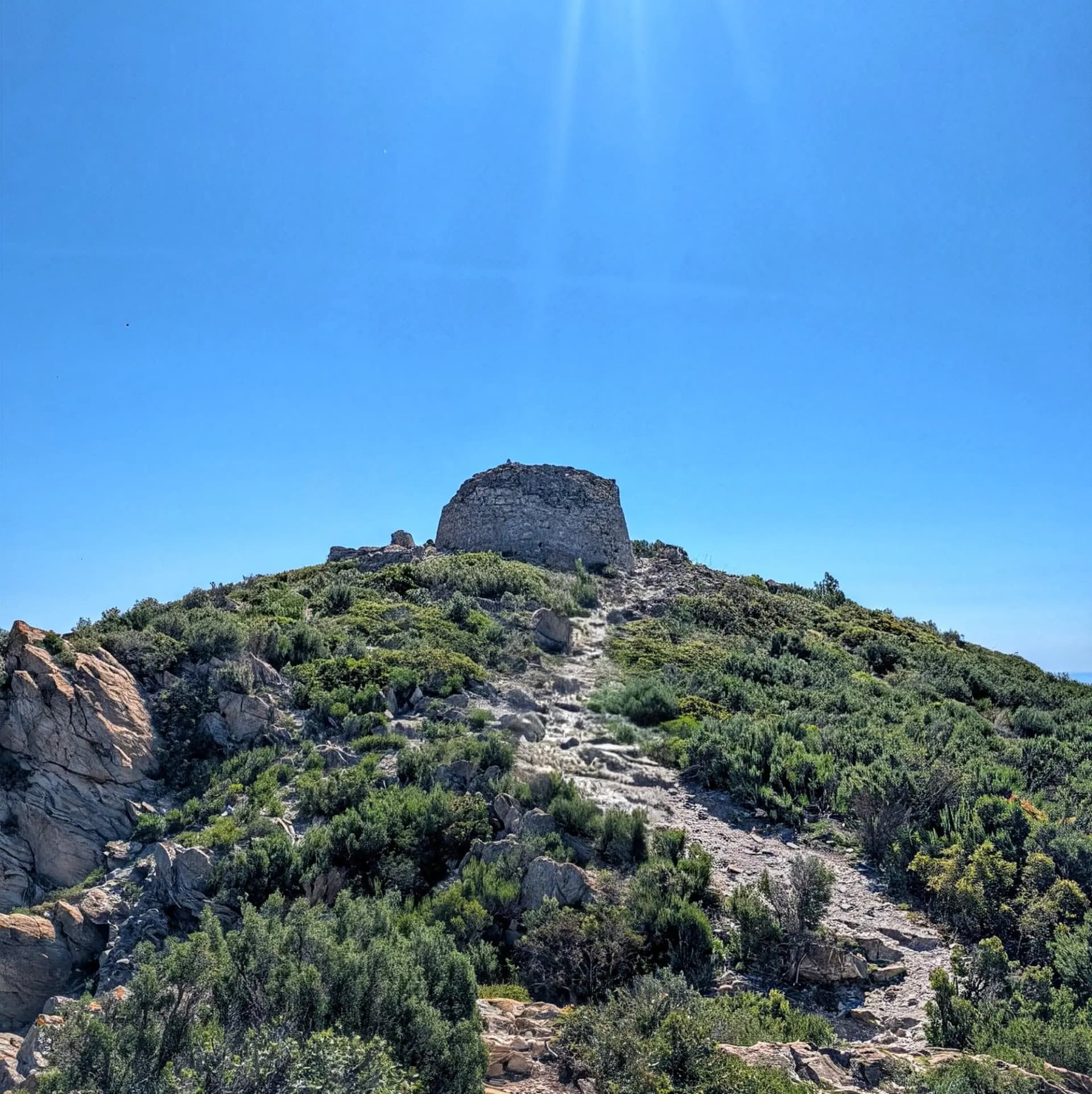The Tower of Norfeu, an ancient stone structure, stands prominently on a hill covered in Mediterranean vegetation, with a rocky path leading up to it under a clear blue sky. Stone tower on a green, rocky hilltop with a winding path under a clear blue sky.