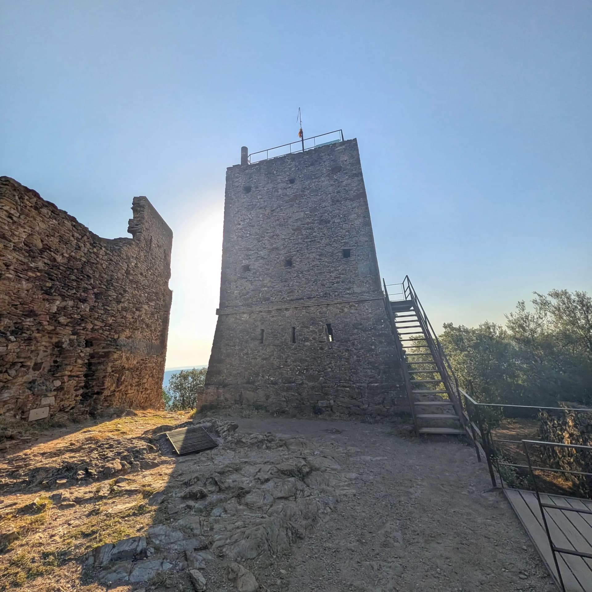 La històrica torre de pedra del Castell de Sant Miquel s'alça contra un cel blau clar, amb vistes a arbres verds i el mar llunyà. Una robusta escala metàl·lica permet l'accés al seu nivell superior. Torre de pedra d'un castell amb escales metàl·liques externes sota cel blau clar. Ruïnes de mur a l'esquerra, arbres i mar al fons.