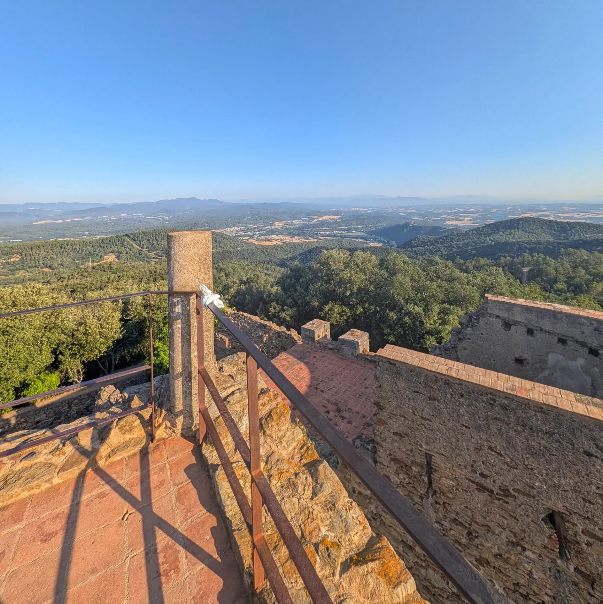 La vista panoràmica des del Castell de Sant Miquel a Girona, amb àmplies vistes dels turons boscosos i les valls llunyanes. Muralla de pedra, terra de rajola i barana de metall emmarquen una vista panoràmica de turons boscosos i valls sota cel blau.