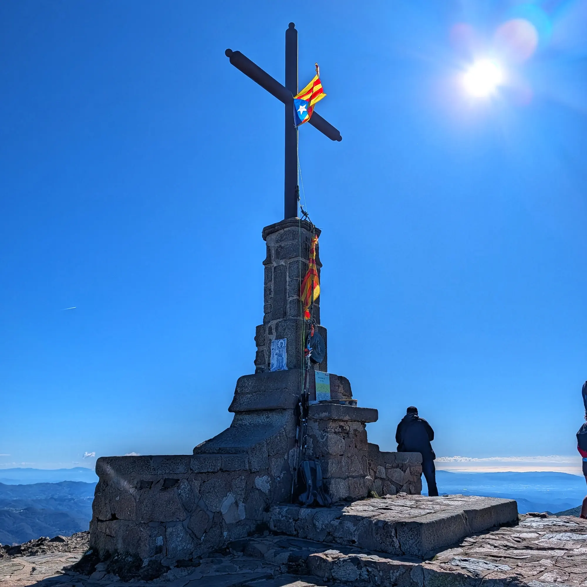 El cim del Matagalls, amb la seva característica creu de pedra i una estelada onejant. Un excursionista gaudeix de les vistes panoràmiques del Parc Natural del Montseny i del mar Mediterrani en la llunyania, sota un cel clar i assolellat. Creu de cim al Matagalls amb estelada, persona contemplant muntanyes i mar llunyà sota cel clar.