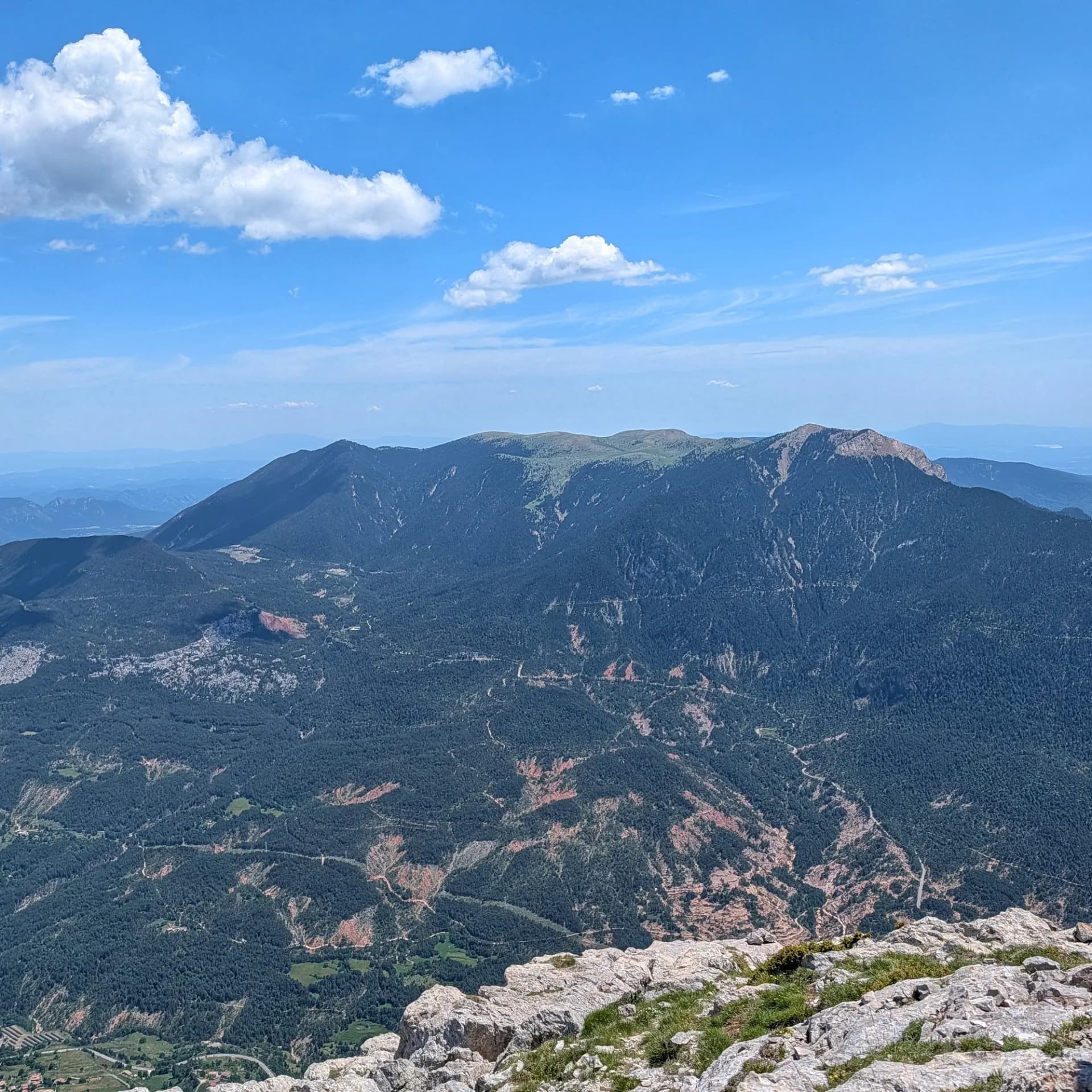 A panoramic view of the Serra d'Ensija and Cap de la Gallina Pelada, captured from the Pollegó Inferior of Pedraforca. The image features a vast mountainous landscape with dense forests and winding mountain roads under a clear blue sky with scattered clouds. Panoramic mountain view from Pedraforca's Pollegó Inferior, showing Serra d'Ensija, Cap Gallina Pelada, forests and blue sky with clouds.