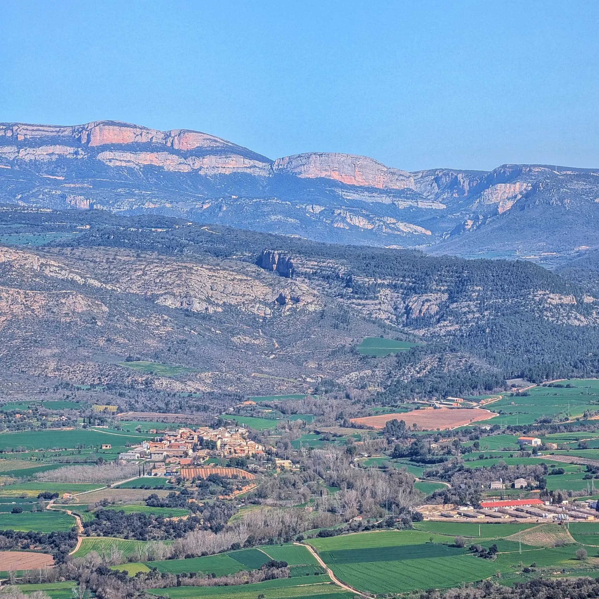 Panoramic view of the impressive Roca Alta, part of the Montsec mountain range, captured from the summit of Munt de Montsonís, showcasing a fertile valley with cultivated fields and a small village. Panoramic view of Roca Alta (Montsec) from Munt de Montsonís, with green valley, fields, and a village.