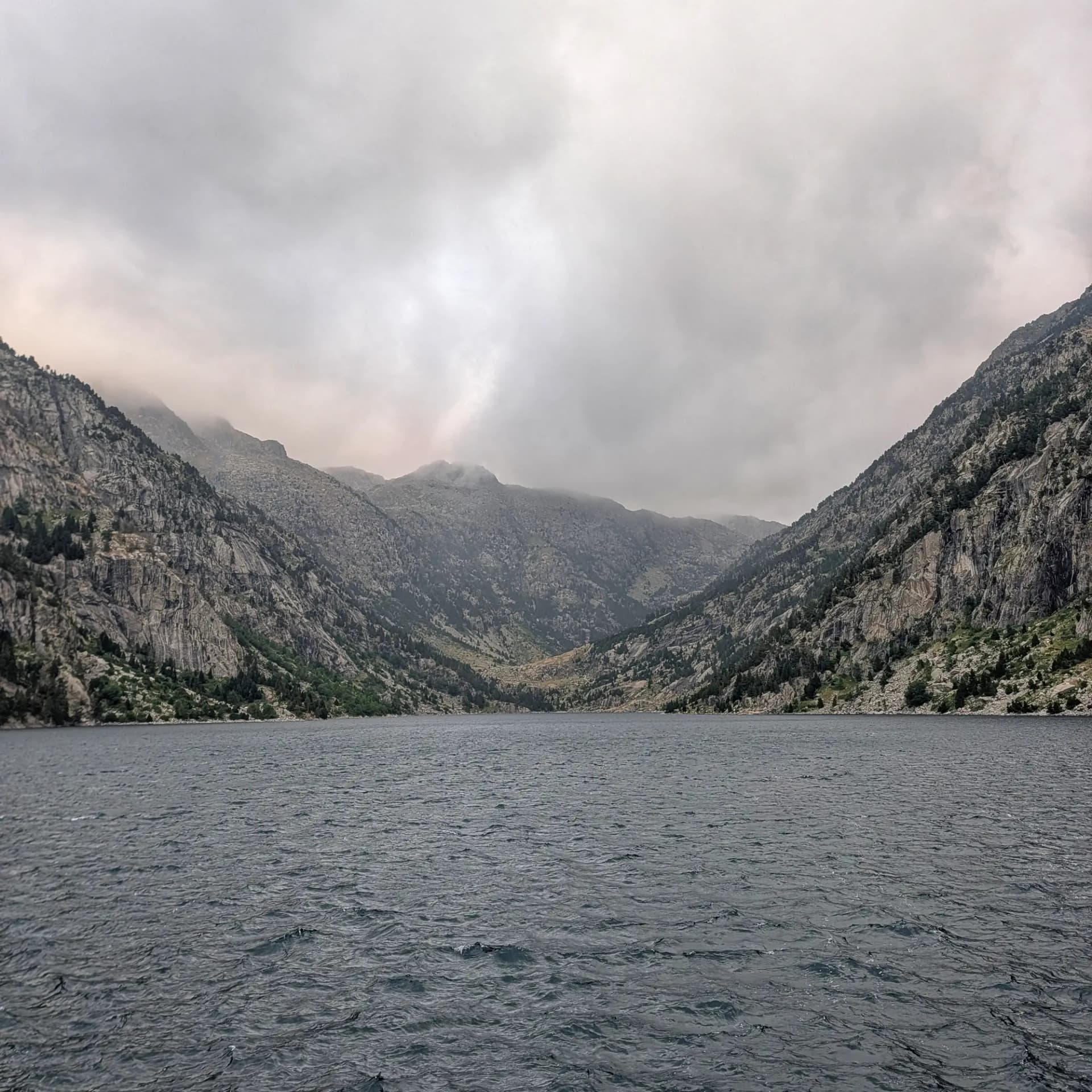 Les aigües de la Presa de Cavallers, punt de partida per a la ruta d'ascensió a cims com el Comaloforno, rodejat de majestuoses muntanyes granítiques sota un cel cobert. Llac fosc i serè envoltat per imponents muntanyes rocoses amb escassa vegetació verda sota un cel ennuvolat i gris.