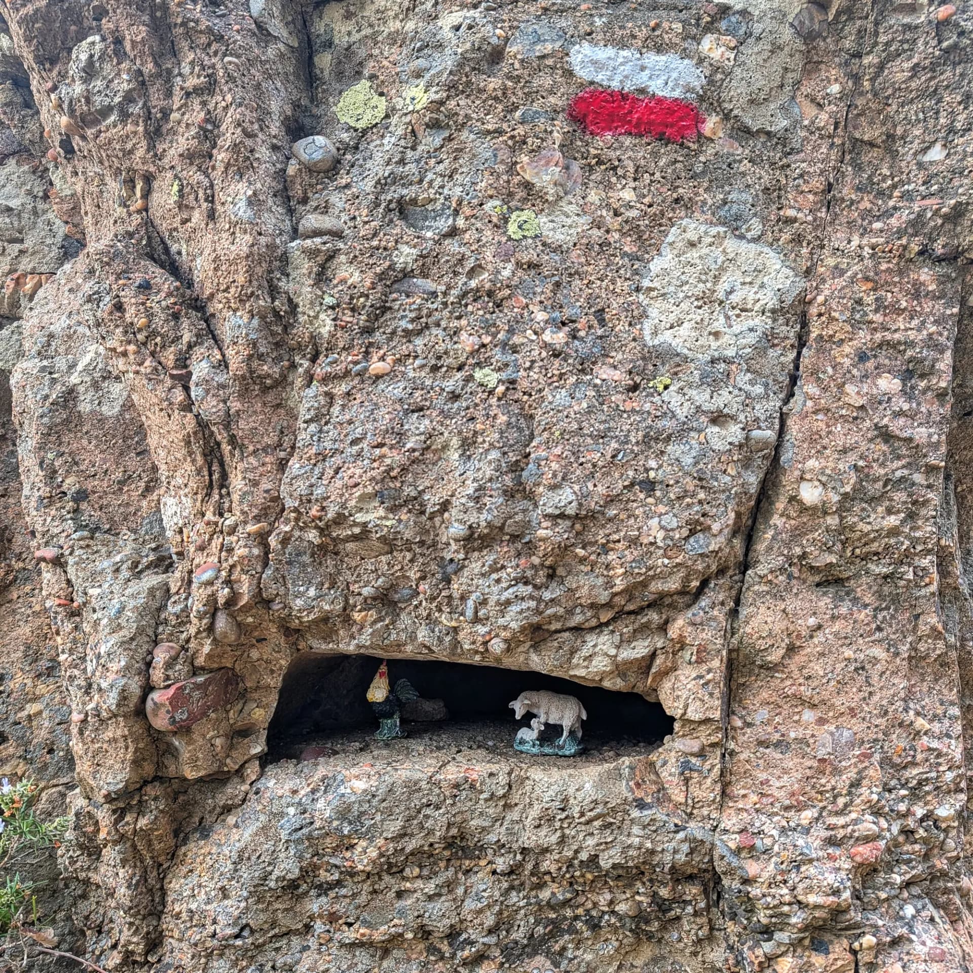 Un detall singular en una paret de roca a Els Estrets, amb senyalització de sender GR i petites figures de gall, ovella i xai en una fornícula, observat durant la tornada del Castell d'Escornalbou. Paret de roca amb senyal de GR i fornícula amb figures de gall, ovella i xai.