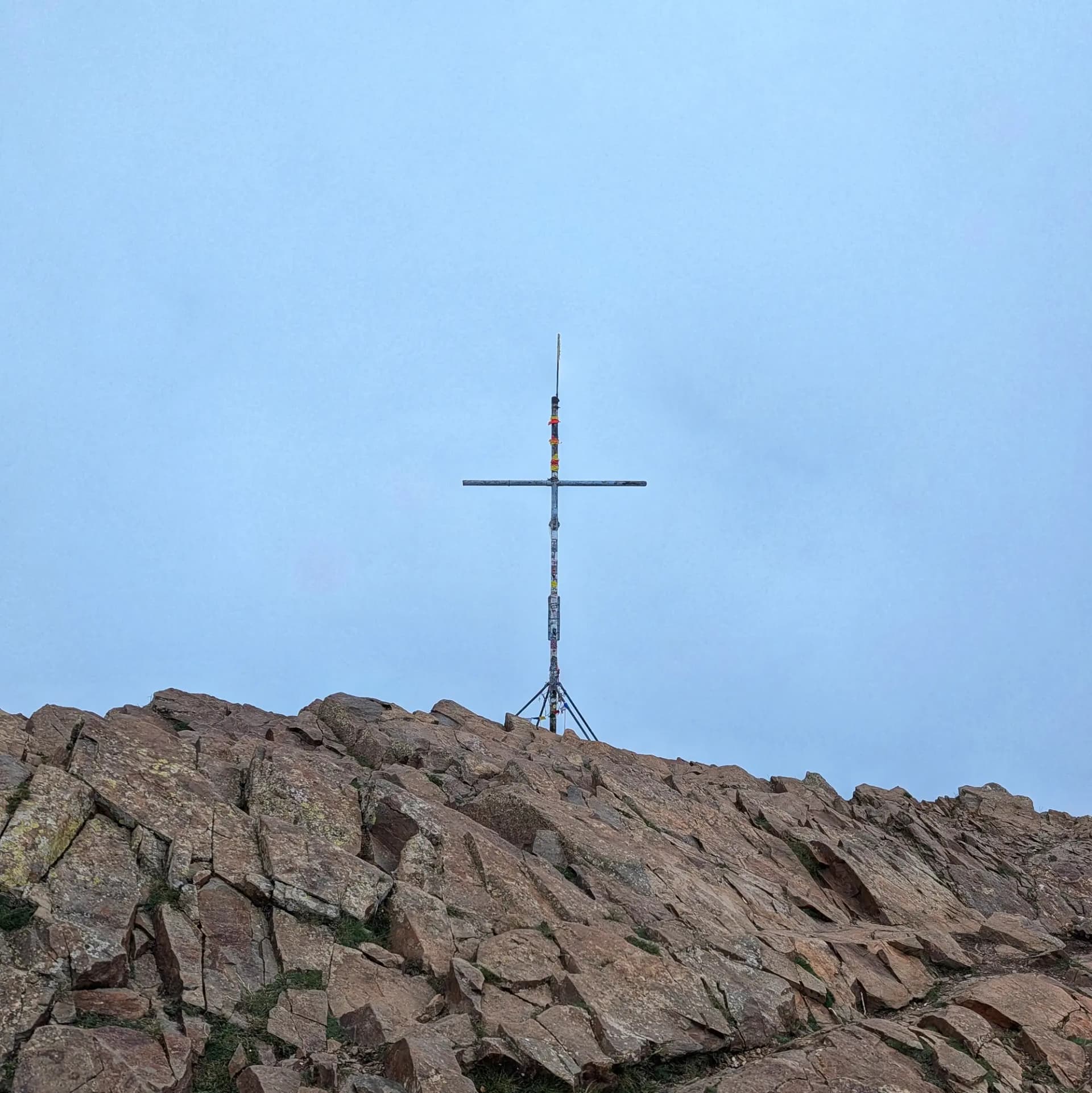 La creu de cim metàl·lica de Les Agudes, situada sobre les característiques roques marró-vermelloses del Massís del Montseny, sota un cel clar. Creu metàl·lica al cim de Les Agudes, sobre roques marró-vermelloses sota un cel clar.