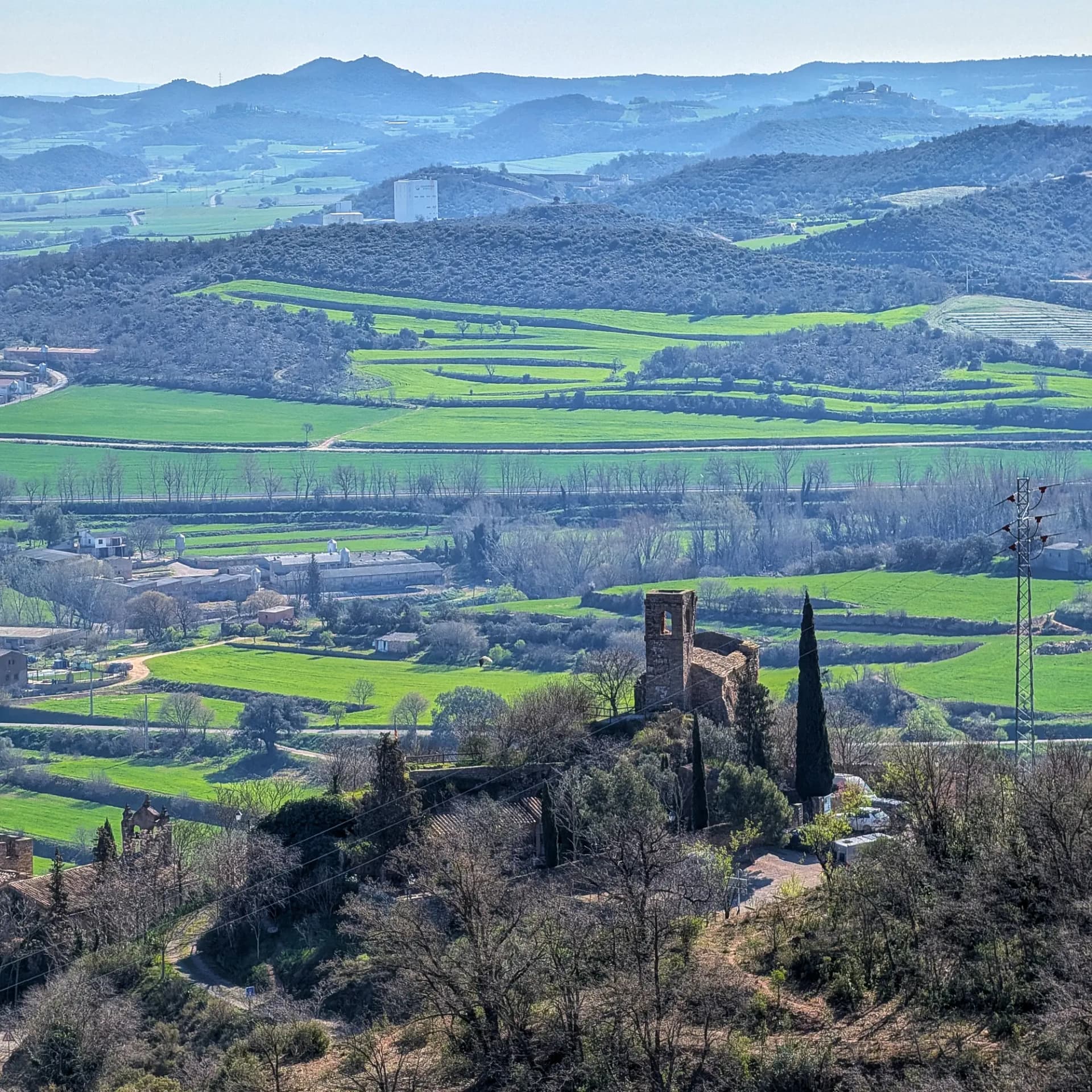 Panoramic view of Montsonís Hermitage and its stone bell tower, surrounded by green fields and hills, taken during the ascent. Stone hermitage of Montsonís with bell tower, surrounded by green fields and hills under blue sky, elevated view.