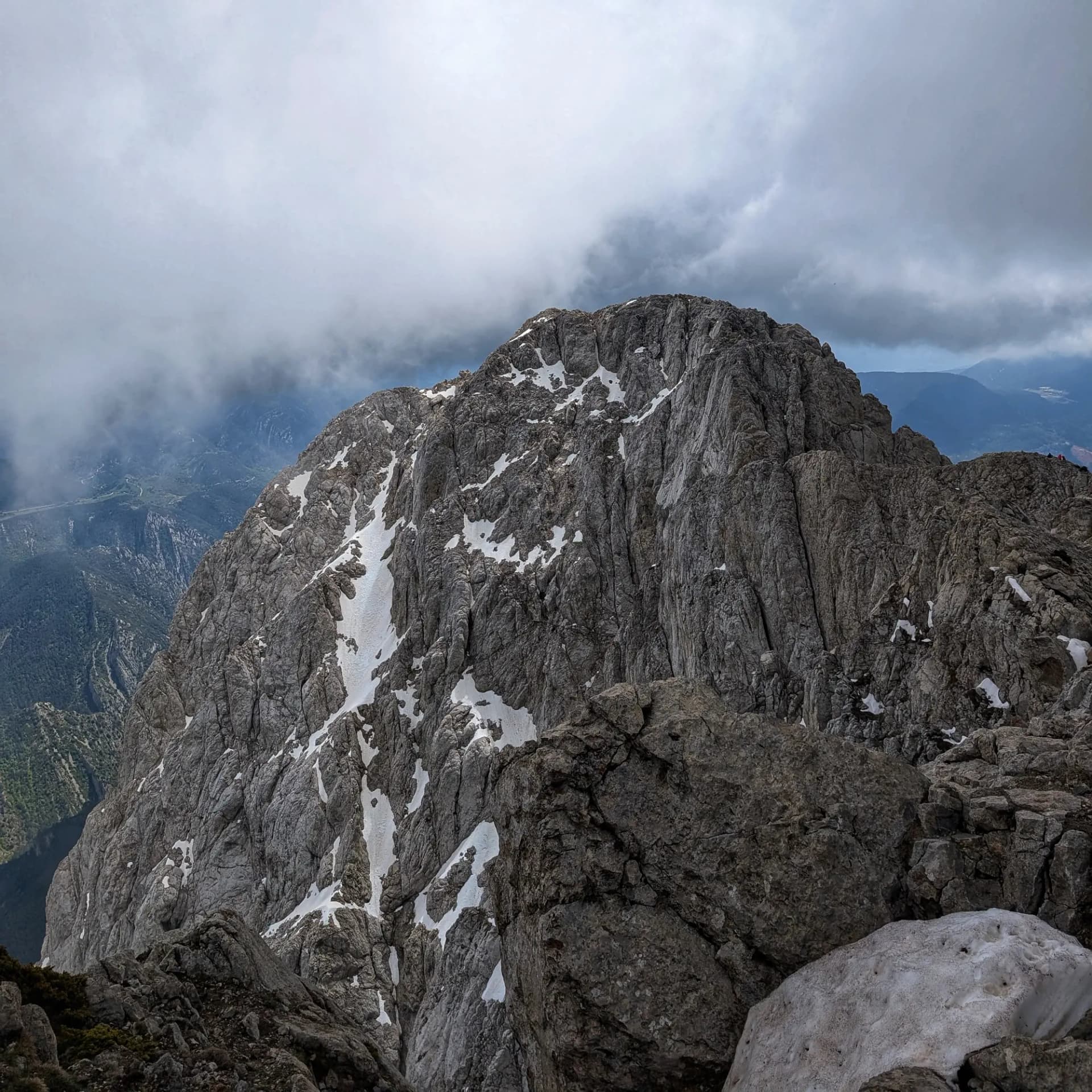 Les imponents parets rocoses del Massís del Pedraforca, amb diverses clapes de neu, fotografiades des del cim del Pollegó Superior del Pedraforca sota un cel parcialment cobert. Pic rocós gris amb clapes de neu sota cel ennuvolat, vist des d'un punt elevat.