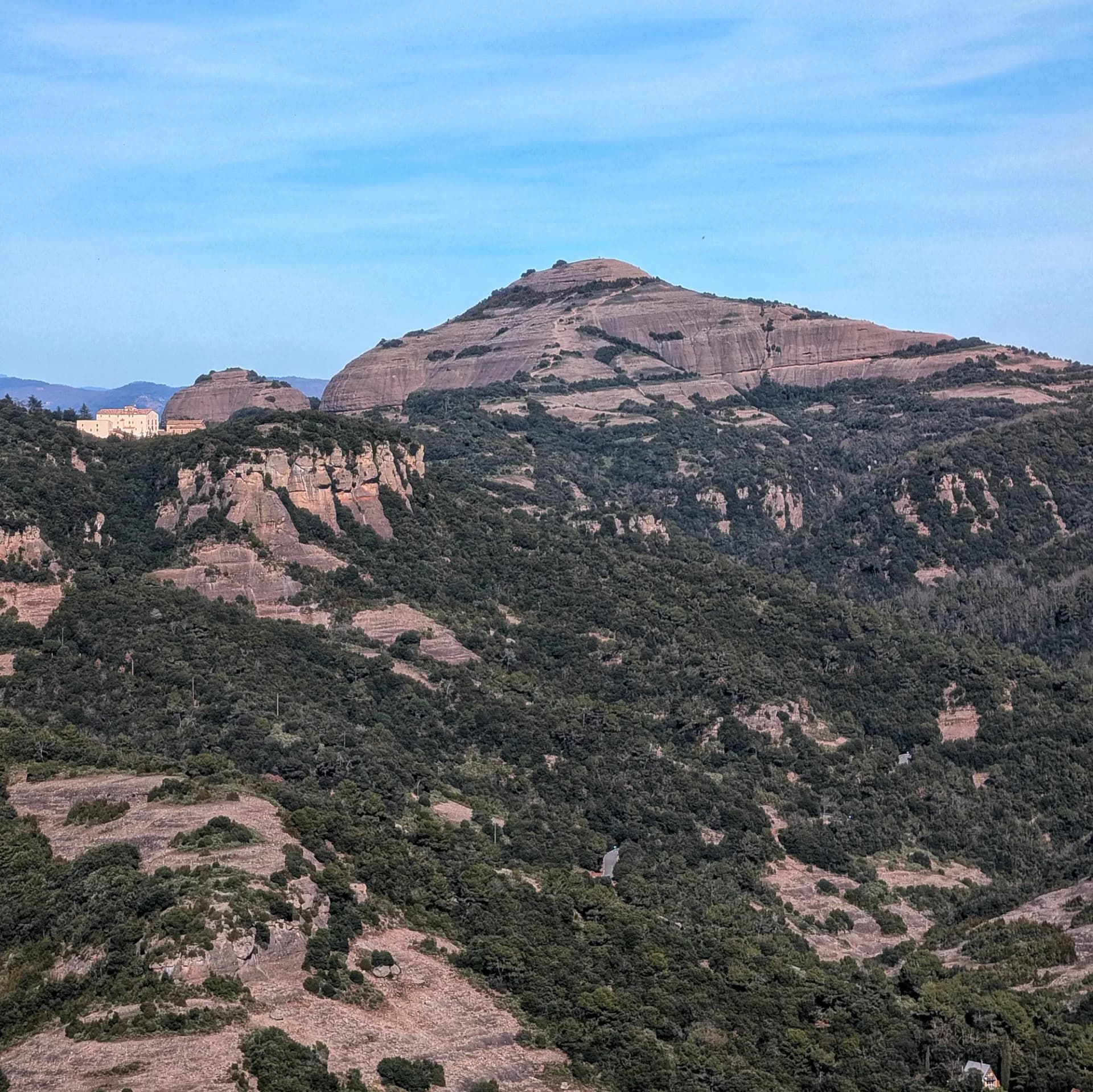 Vista panorámica del Montcau, una de las cumbres emblemáticas del Parque Natural de Sant Llorenç del Munt i l'Obac. La imagen muestra sus características formaciones rocosas estratificadas, la densa vegetación mediterránea y una edificación en un cerro vecino, capturada durante el ascenso a La Mola. Vista del Montcau con sus capas de roca y denso bosque verde, observado durante una ruta en el Parque Natural de Sant Llorenç del Munt i l'Obac.