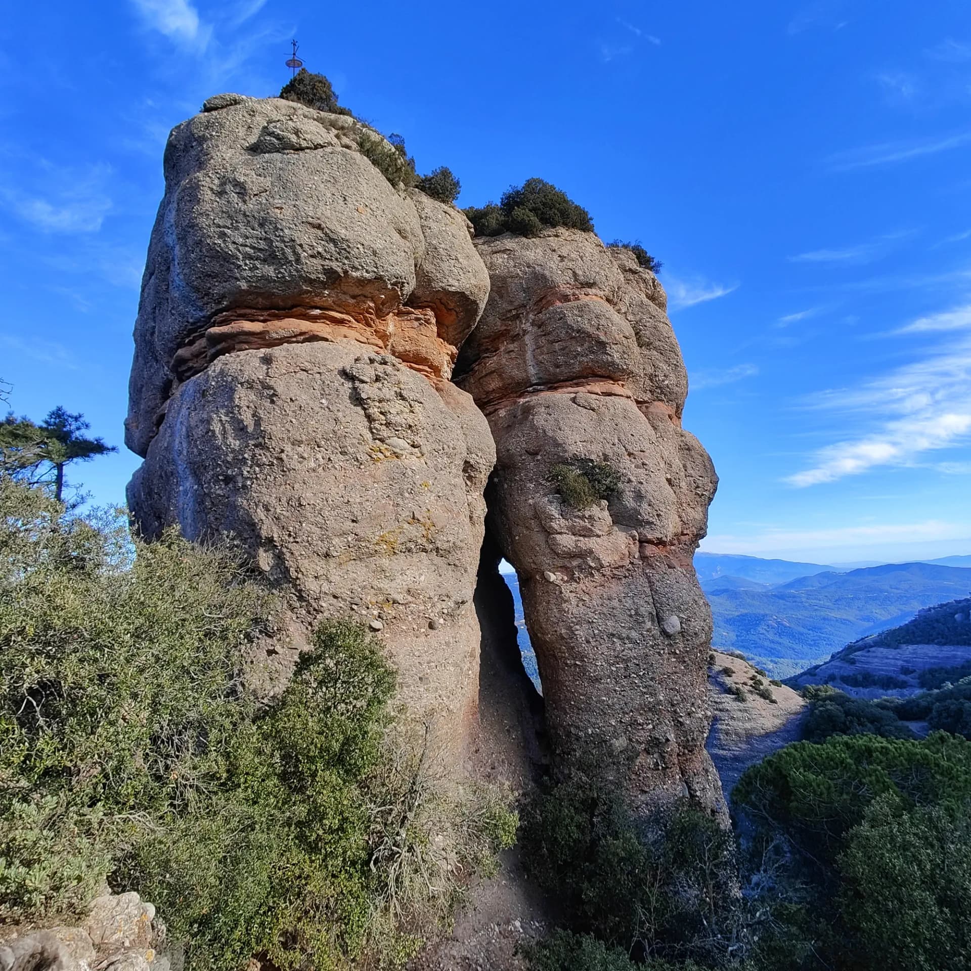 La distintiva formació rocosa del Morral del Drac, a la serra de Sant Llorenç del Munt. Dues formacions rocoses imponents amb una estreta escletxa, vegetació esparsa i creu al cim, cel blau.