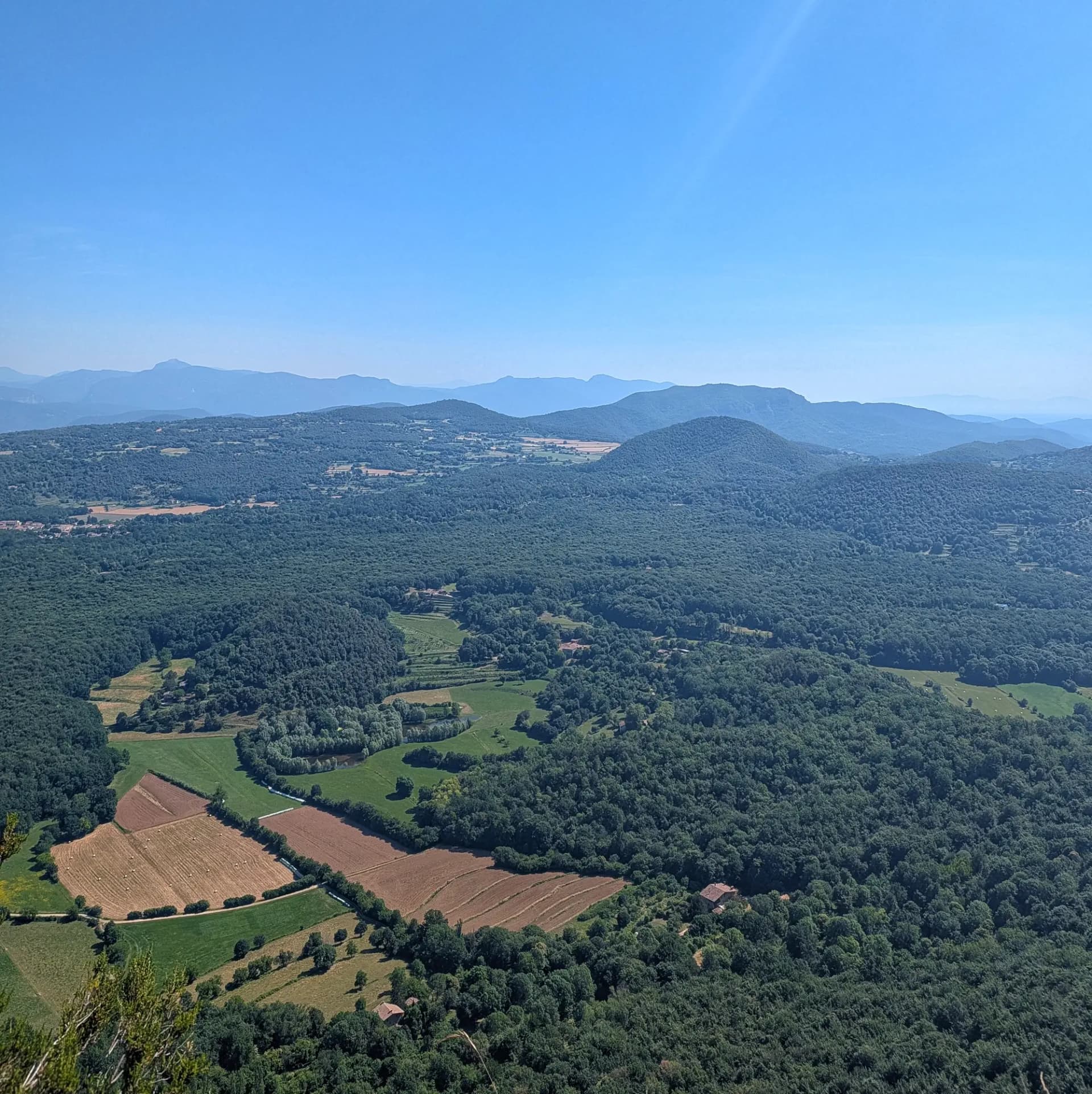 Impresionante vista panorámica de la exuberante región de la Garrotxa, mostrando sus densos bosques, el mosaico de campos y colinas, vistas desde la cumbre de Roca Lladre. Vista panorámica de densos bosques verdes, campos cultivados, casas dispersas y montañas lejanas bajo un cielo azul.