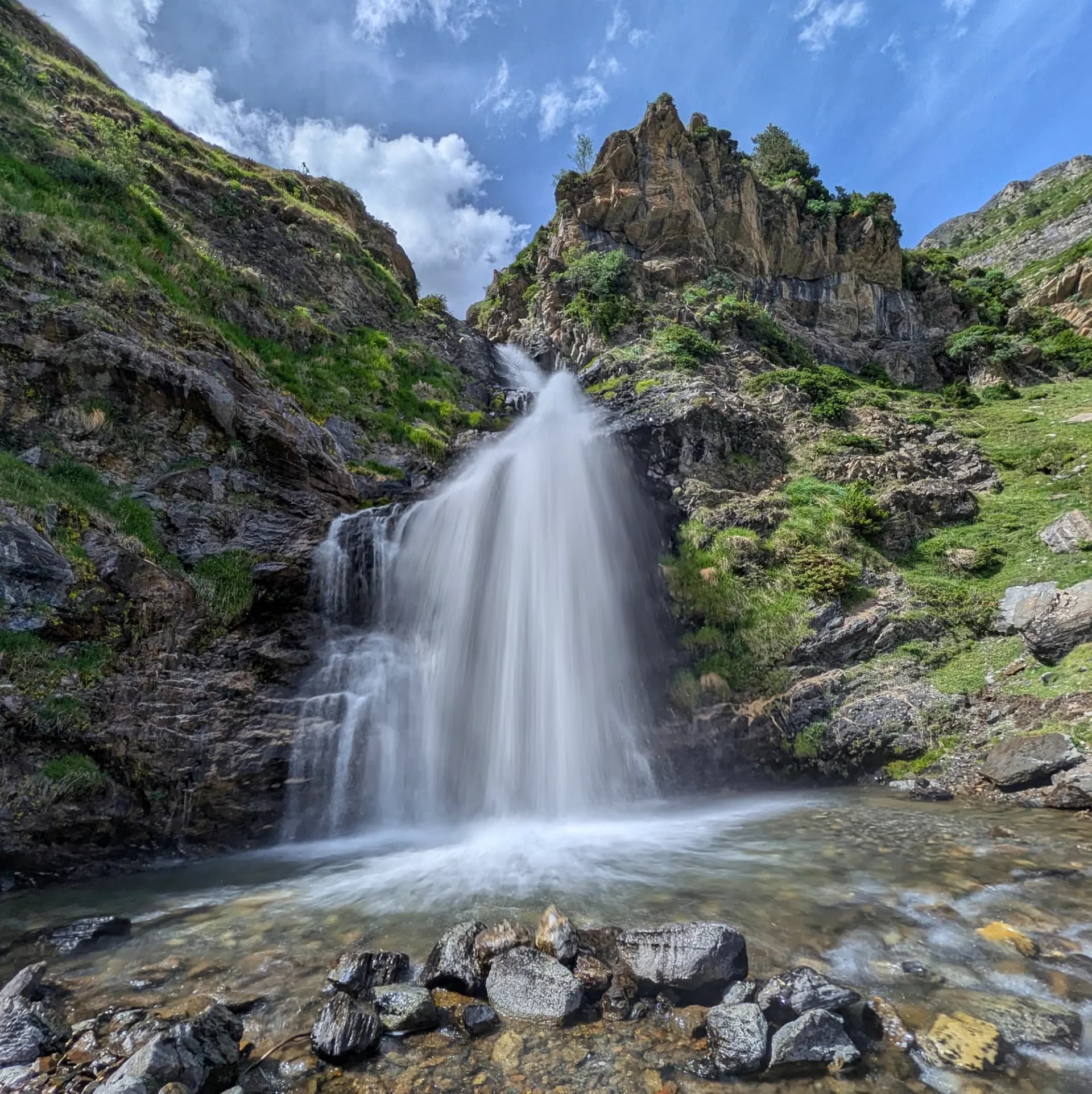 La majestuosa Cascada dels Prats, un salt d'aigua potent que flueix per penya-segats escarpats i vessants verds. A la ruta cap als ibones de Barbarisa des del Collado de Sahún. Imatge de llarga exposició d'una cascada potent caient per una paret de muntanya rocosa i verda sota cel blau.