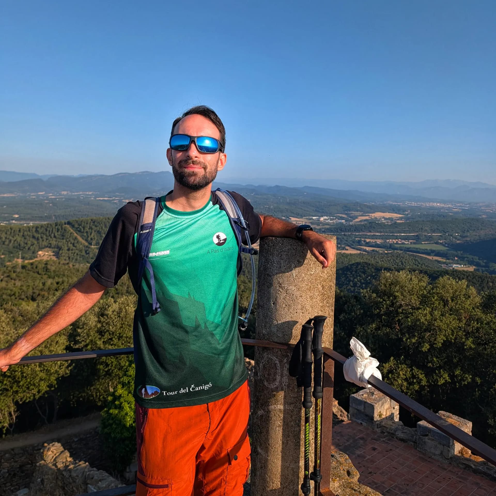 Castell de Sant Miquel Man in sunglasses and hiking gear on a viewpoint, smiling and overlooking a vast green landscape with distant mountains.