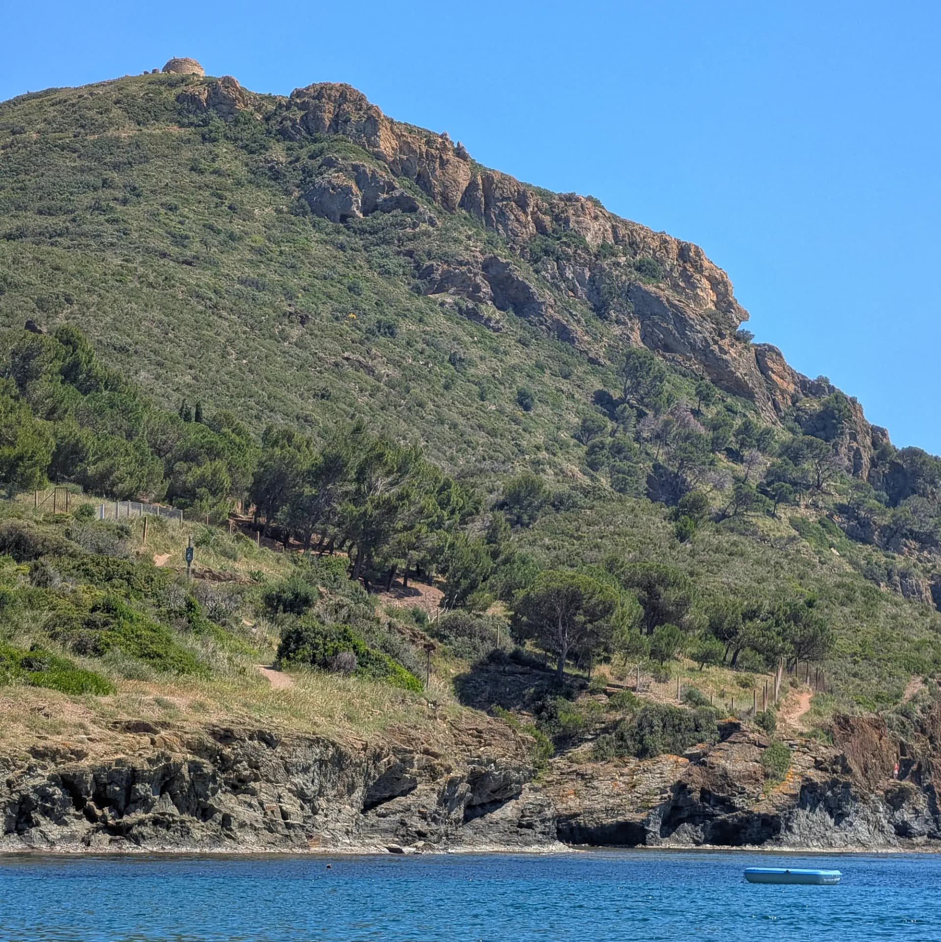 The Torre de Norfeu, an ancient coastal watchtower, stands atop a verdant, rocky hillside. The image is captured from the waters of Cala Pelosa, with a small floating platform visible in the clear blue sea. Historic tower on a rocky, green hillside overlooking blue Mediterranean sea with a floating platform.