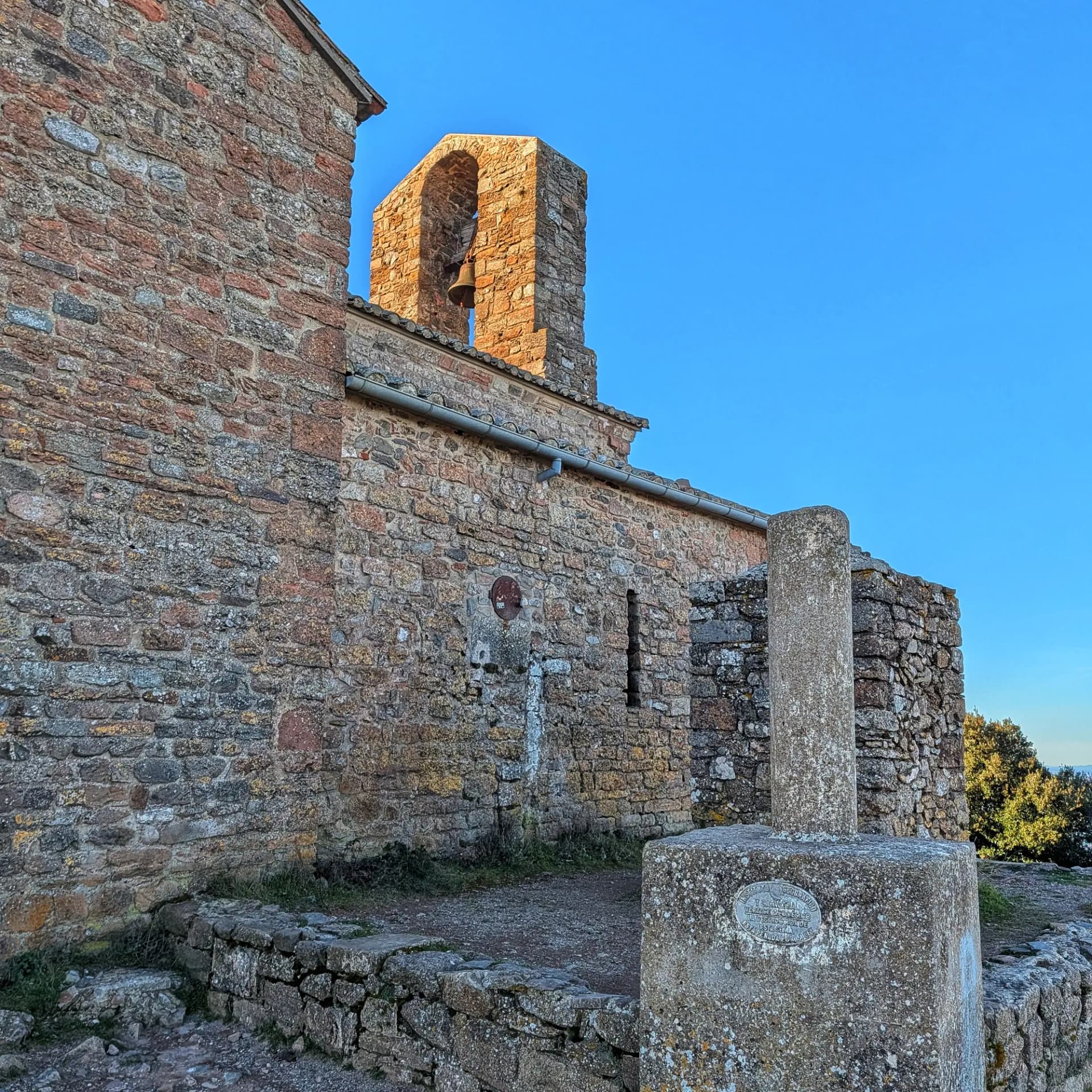 Vèrtex geodèsic de la Mola de Sant Llorenç, situat al costat del monestir romànic, durant una ruta pel Parc Natural de Sant Llorenç del Munt i l'Obac amb ascensió a La Mola. Edifici de pedra antic amb campanar de paret i vèrtex geodèsic sota un cel blau clar.